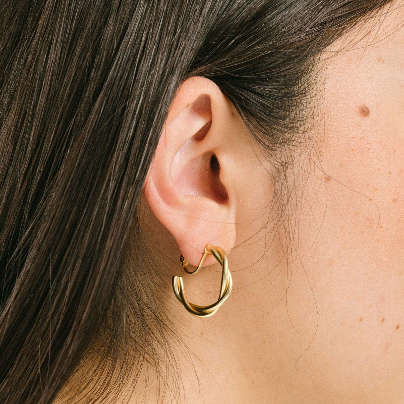 A model wearing the Twist Hoop Earrings feature a secure and comfortable screwback clip-on closure ideal for all ear types. Wear these earrings for up to 8-12 hours per day with a secure hold and the ability to be manually adjusted to the individual's size. Crafted from gold tone plated copper alloy, these earrings come is a single pair.
