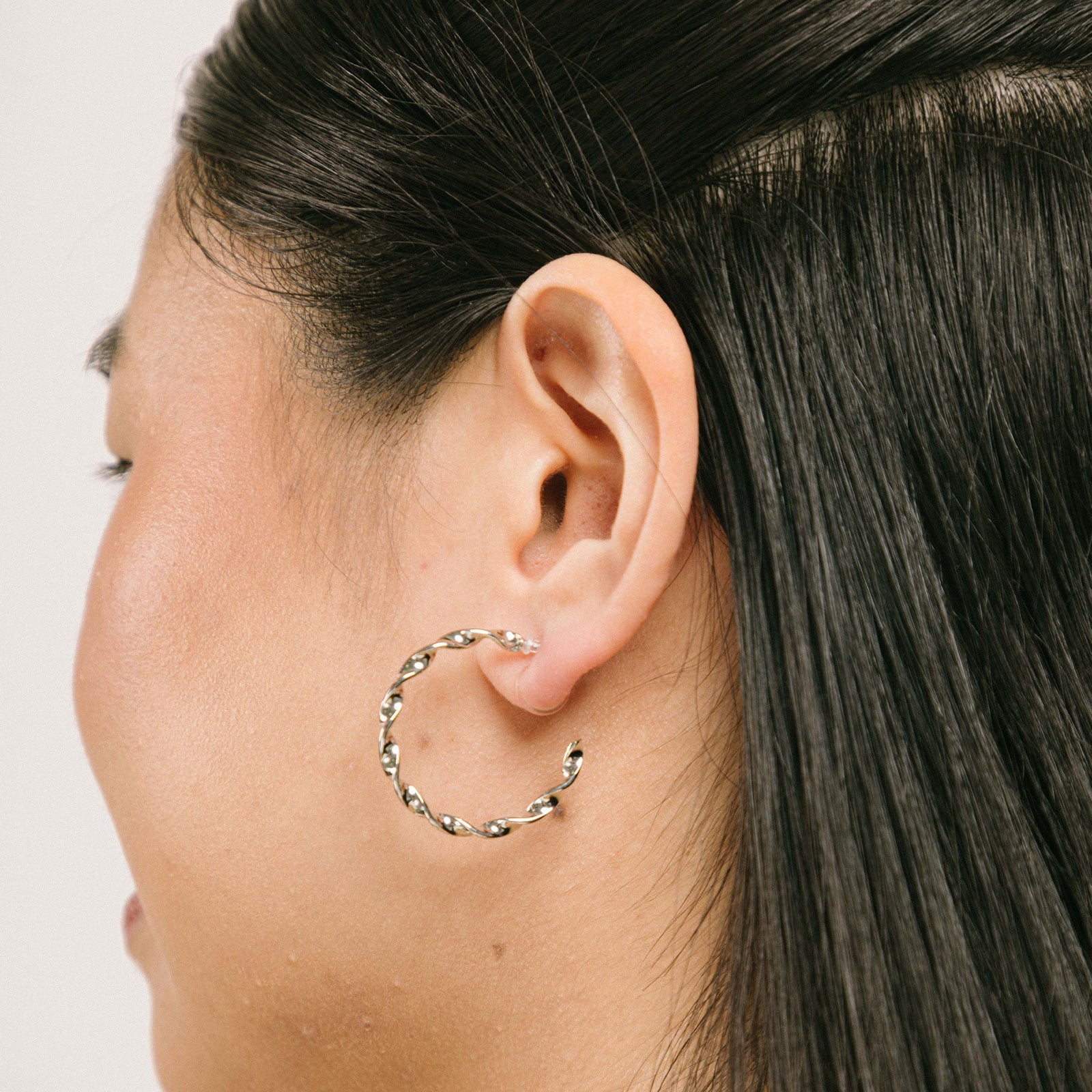 A model wearing the Silver Wave Hoop Clip-On Earrings are perfect for all ear types. Whether you have thick or large ears, or sensitive, small or thin ears, these earrings offer a medium-secure hold and can be comfortably worn for 8-12 hours. The earrings are made of silver-tone metal alloy and offer no ability to adjust - each purchase is for one pair. You can also find these earrings in a gold version.