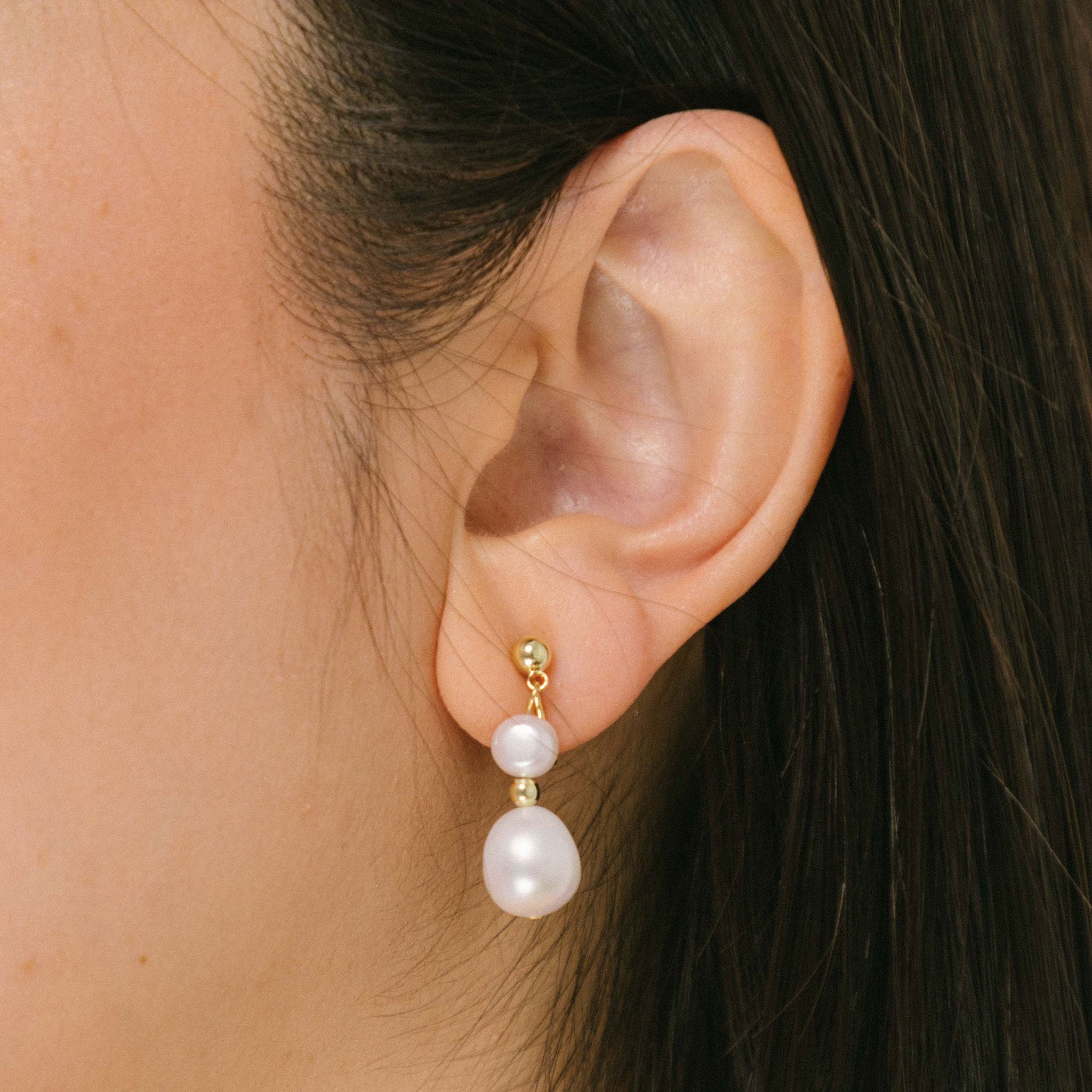 A model wearing the Duo Pearl Clip-On Earrings in Gold feature a mosquito coil clip-on closure ideal for all ear types. Enjoy medium secure hold and comfortable wear for up to 24 hours - gently squeezing the padding forward once on the ear for adjustable fit. Crafted with freshwater pearls and 18K gold plated stainless steel, each piece features unique natural variations in size and colour.