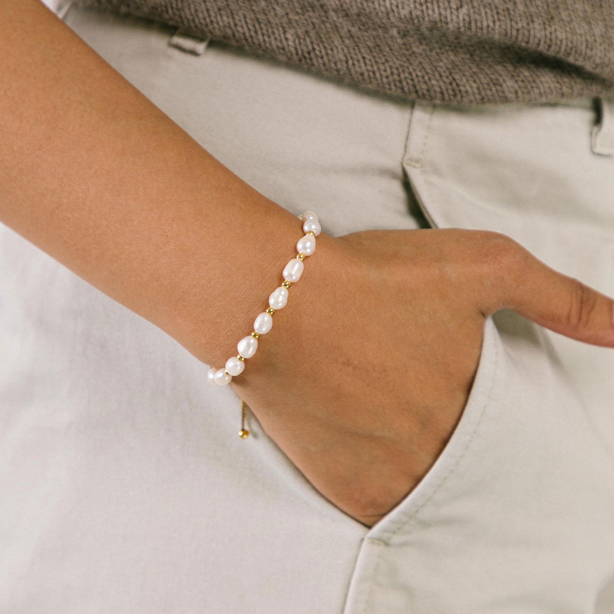 A model wearing the Emi Pearl Bracelet is crafted from 18K Gold Plated Stainless Steel and Freshwater Pearls, allowing for a small degree of variation in size and hue. The bracelet is water-resistant and hypoallergenic.