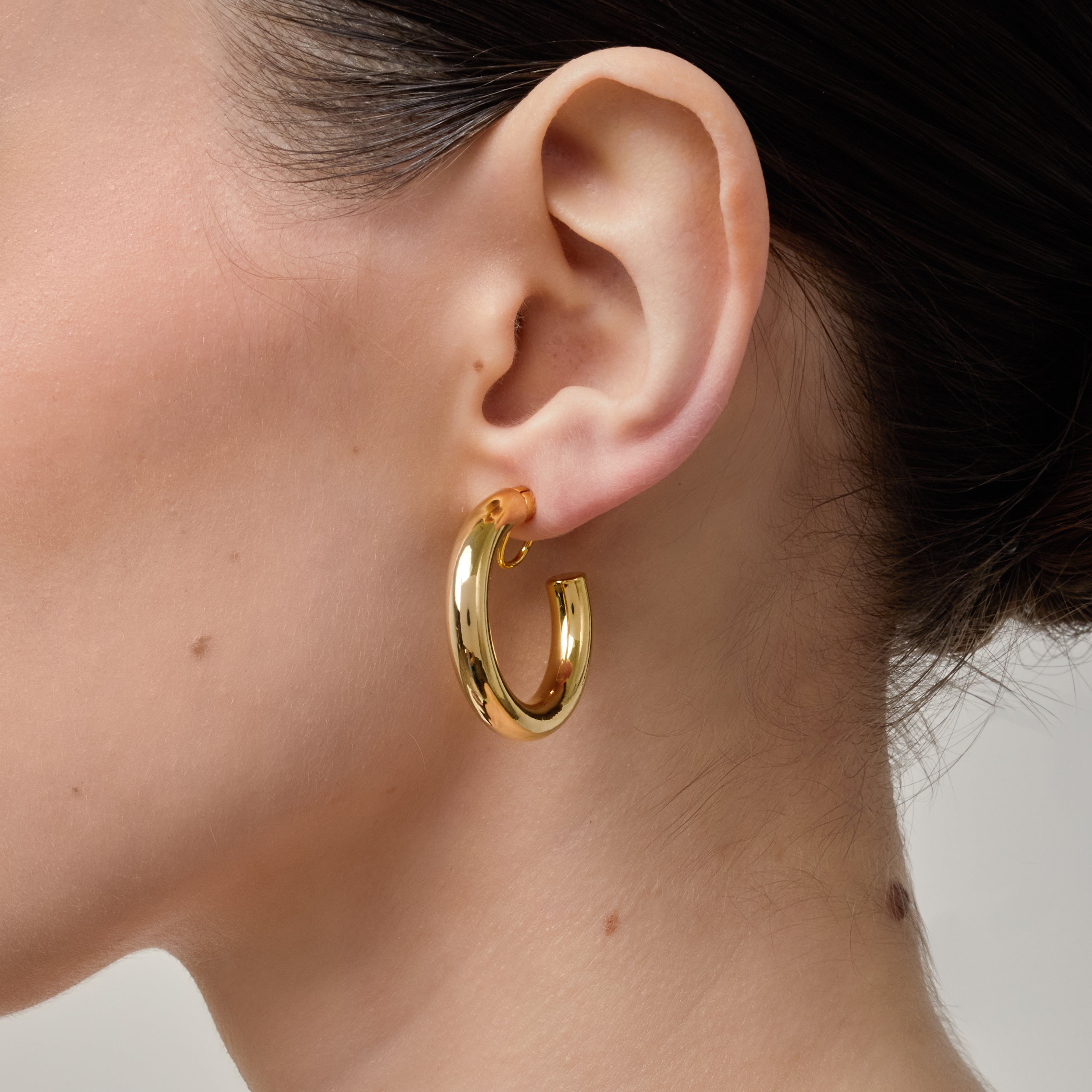 A model wearing the Allie Hoop Clip On Earrings in Gold, which are perfect for all ear types. The Mosquito Coil Clip-On Closure is suitable for Thick/Large, Sensitive, Small/Thin, Stretched/Healing, and Keloid Prone Ears. Made of a Gold tone copper alloy, each purchase includes one pair.