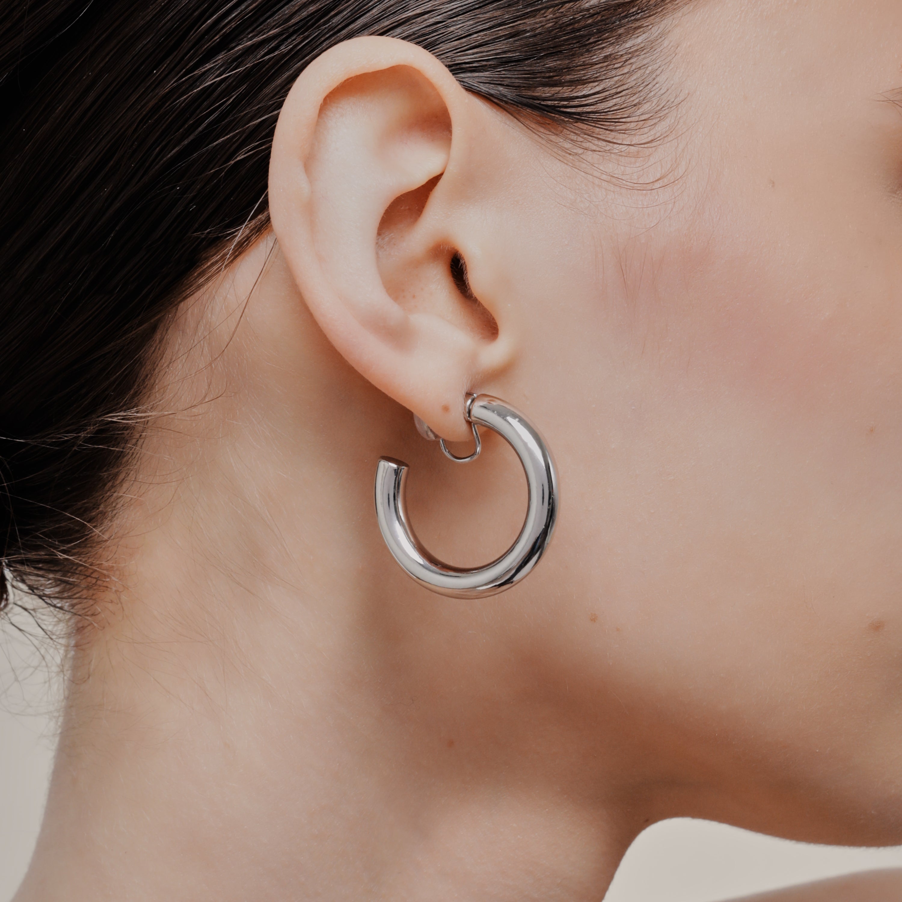A model wearing the Allie Hoop Clip On Earrings in Silver, which are perfect for all ear types. The Mosquito Coil Clip-On Closure is suitable for Thick/Large, Sensitive, Small/Thin, Stretched/Healing, and Keloid Prone Ears. Made of a silver tone copper alloy, each purchase includes one pair.