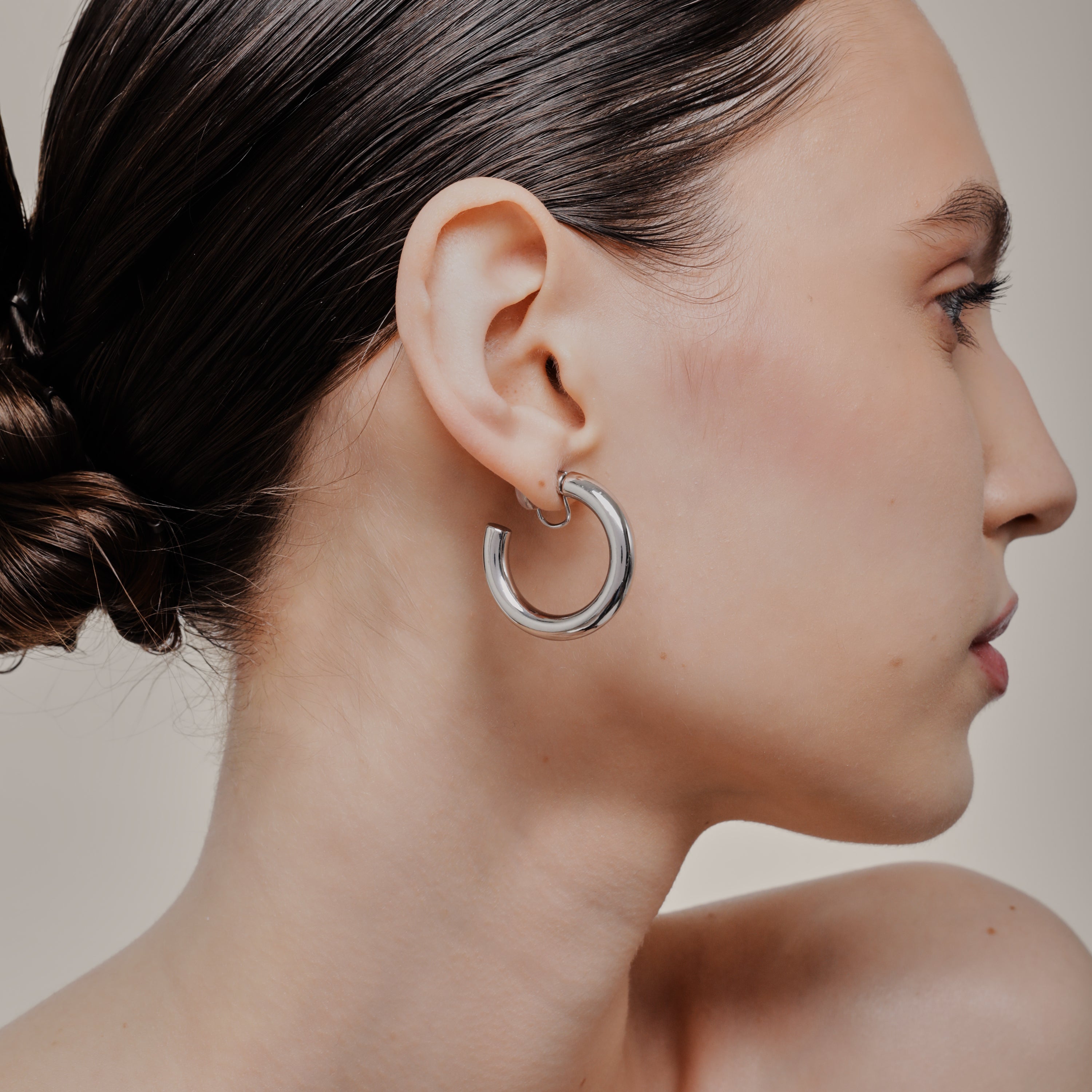 A model wearing the Allie Hoop Clip On Earrings in Silver, which are perfect for all ear types. The Mosquito Coil Clip-On Closure is suitable for Thick/Large, Sensitive, Small/Thin, Stretched/Healing, and Keloid Prone Ears. Made of a silver tone copper alloy, each purchase includes one pair.