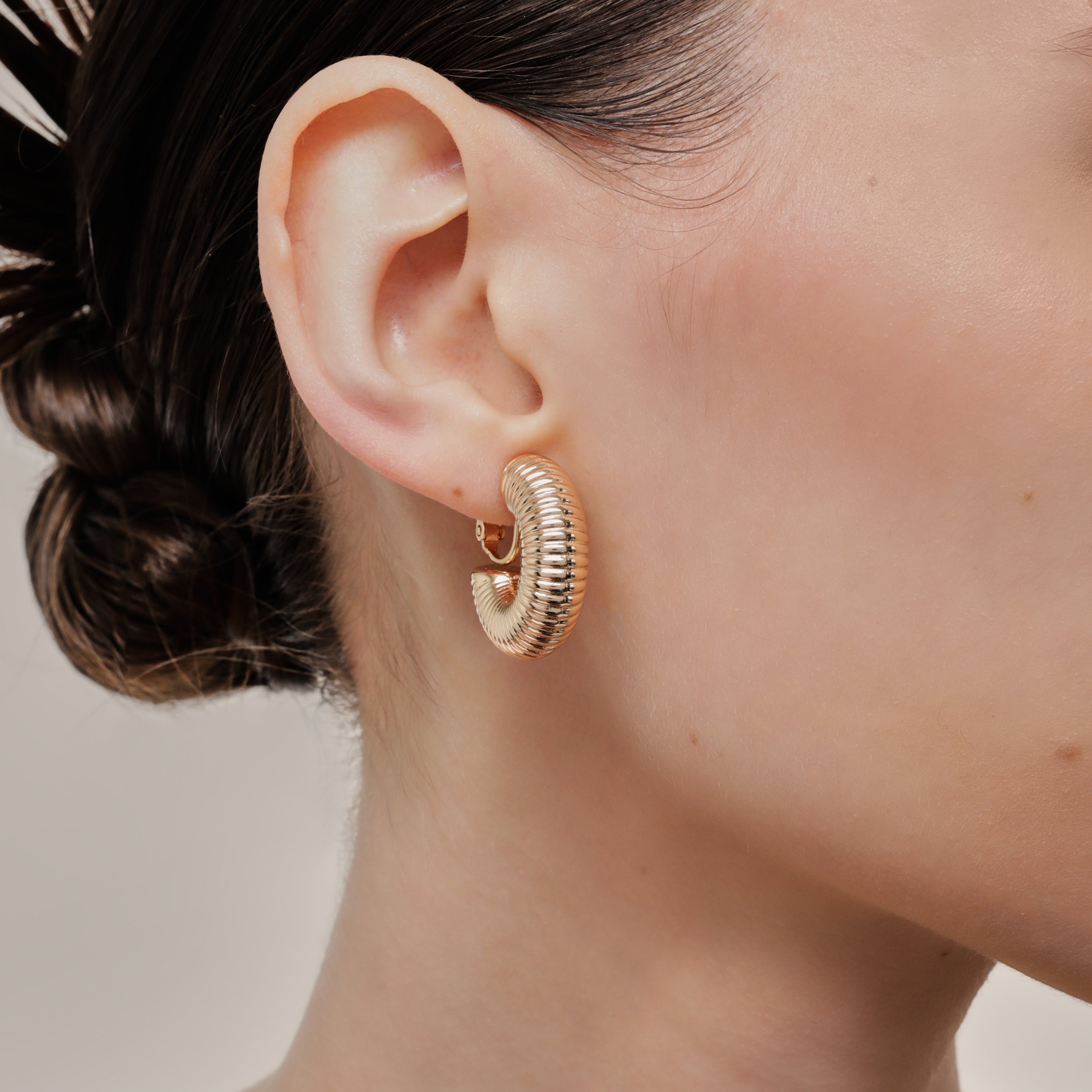 A model wearing the Bold Ribbed Hoop Clip On Earrings in Gold. Our screwback closure and manual adjustment capabilities cater to a wide range of needs, including thick, sensitive, small, and keloid-prone ears. Easily wear them for 8-12 hours for all-day fashion. Sold in pairs.