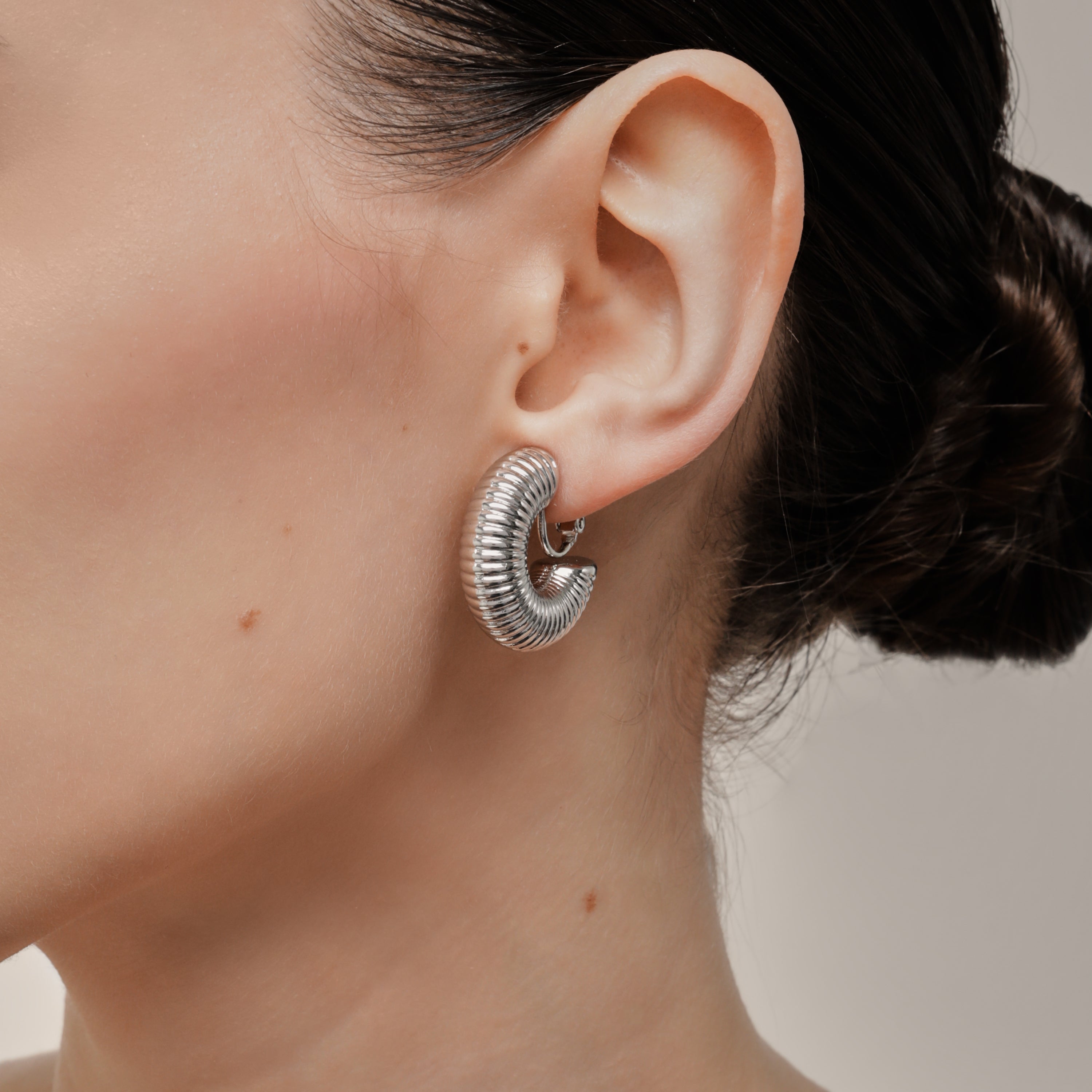 A model wearing the Bold Ribbed Hoop Clip On Earrings in Silver. Our screwback closure and manual adjustment capabilities cater to a wide range of needs, including thick, sensitive, small, and keloid-prone ears. Easily wear them for 8-12 hours for all-day fashion. Sold in pairs.