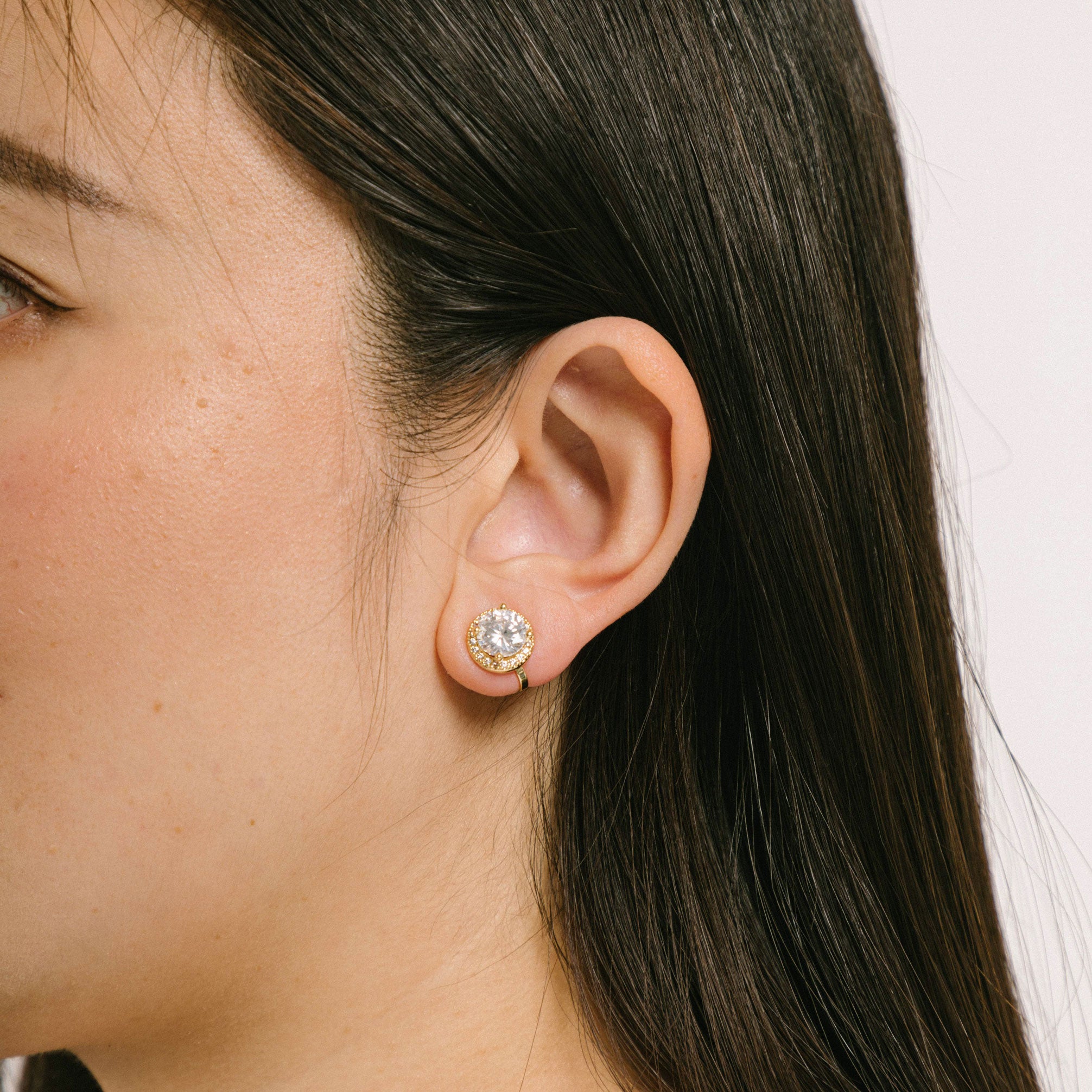 A model wearing the Harper Stud Clip On Earrings in Gold are ideal for any ear type, offering a secure hold for up to 12 hours of comfortable wear. Crafted from gold tone copper alloy and Cubic Zirconia, the earrings feature a padded clip-on closure and removable rubber padding. Please note the item is for one pair only.