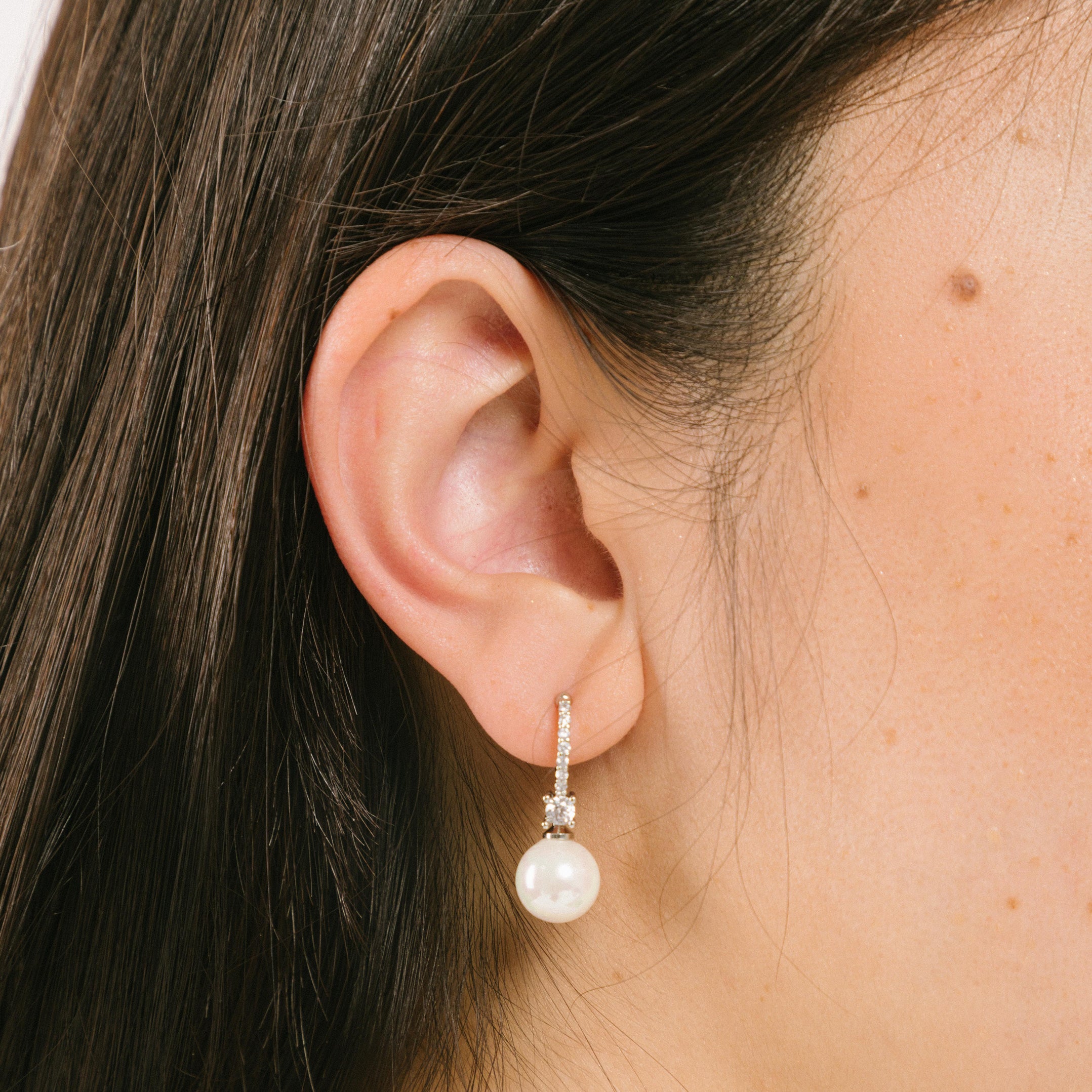 A model wearing the Claire Pearl Clip On Earrings in Silver present a mosquito coil clip-on closure crafted to accommodate all ear types—from thick/large to sensitive, from small/thin to stretched/healing—ensuring a comfortable fit. Intricately constructed from silver tone copper alloy, Cubic Zirconia, and Simulated Faux Pearl, these earrings add a sophisticated touch to any ensemble.