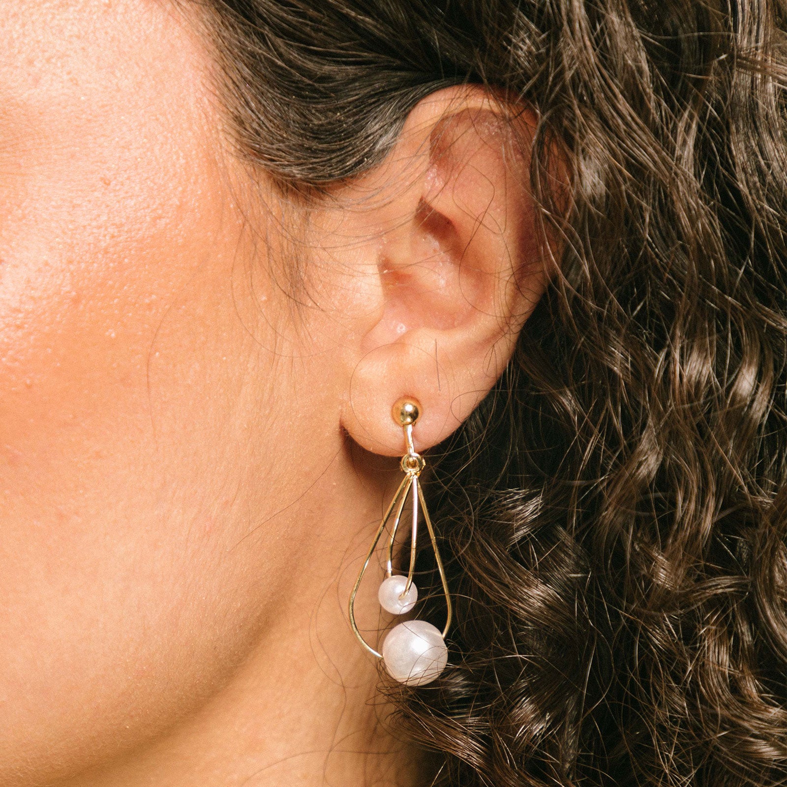 A model wearing Camilla Clip-On Earrings, these alluring screwback clip-on earrings boast a gleaming gold-plated metal alloy and faux pearl, rendering them an exquisite accessory for all ear types. Synergizing with a variety of looks, these figure-eight hoop earrings are ideal for thick/large, sensitive, small/thin, and stretched/healing ears, making them a must-have for any chic ensemble.
