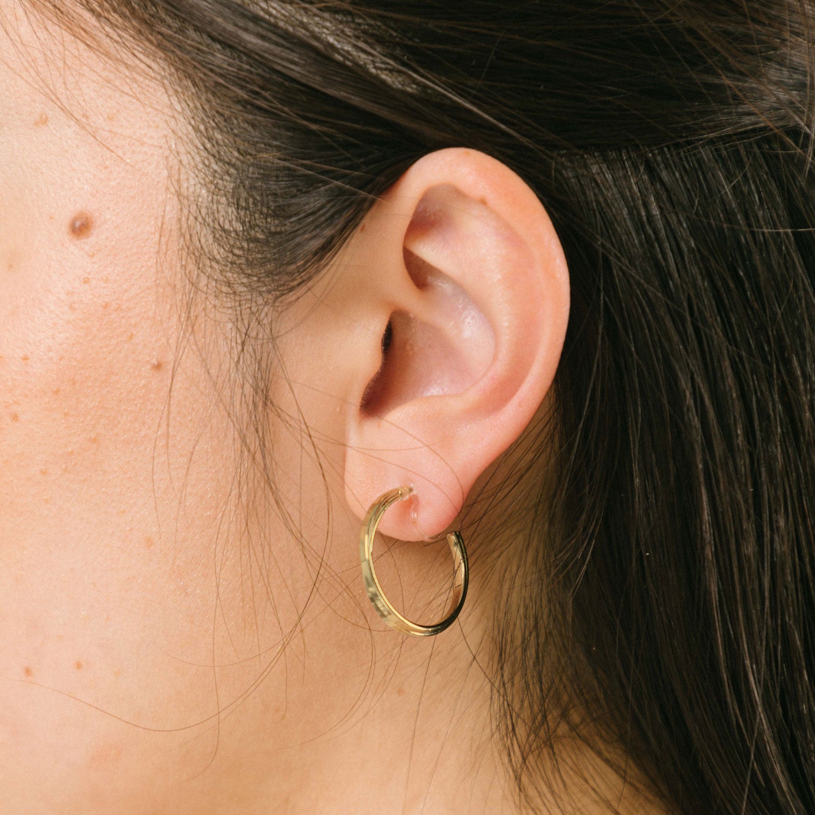 A model wearing the Gold Cassie Hoop Clip-On Earrings feature a resin closure, making them ideal for all ear types, including thick/large ears, sensitive ears, small/thin ears, and stretched/healing ears. On average, pairs are comfortably worn for 8-12 hours with a medium secure hold, and they are not adjustable. This item is sold as a single pair and is also available in Silver. The earrings are constructed with gold-tone metal alloy.