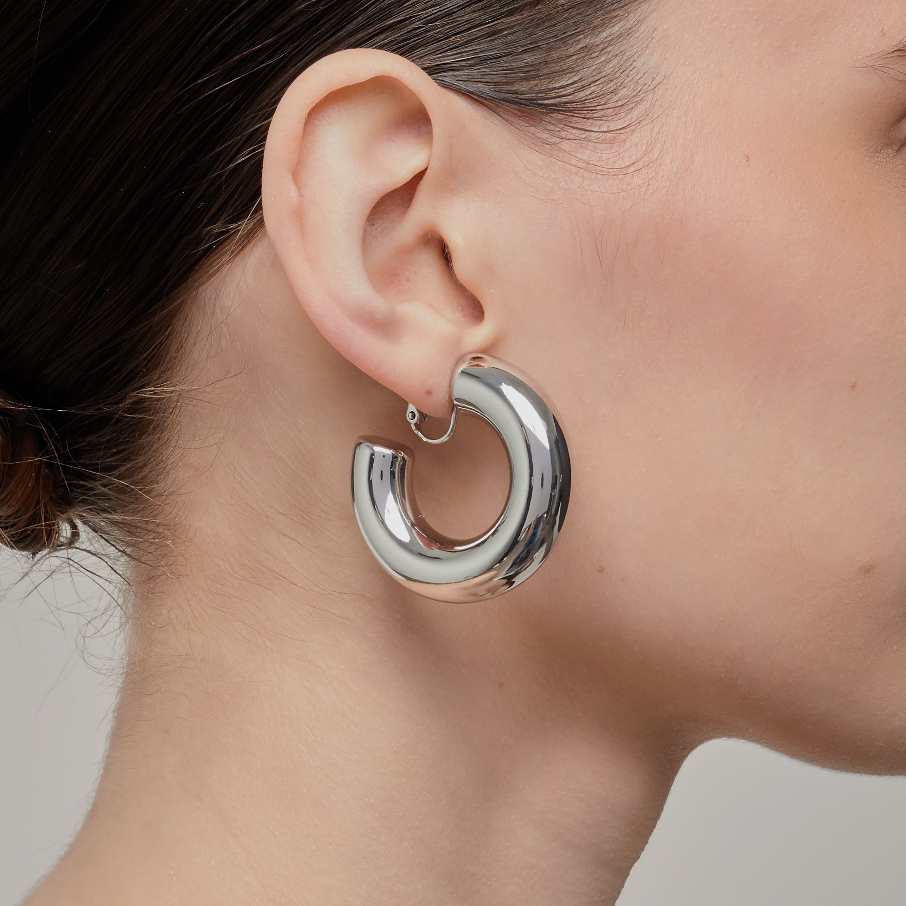A model wearing the Chunky Hoop Clip On Earrings in Silver. With a secure screwback closure, these earrings are suitable for a variety of ear sizes, including sensitive and keloid-prone ears. Designed for long-lasting wear and can be manually adjusted for a perfect fit. Note: includes one pair.