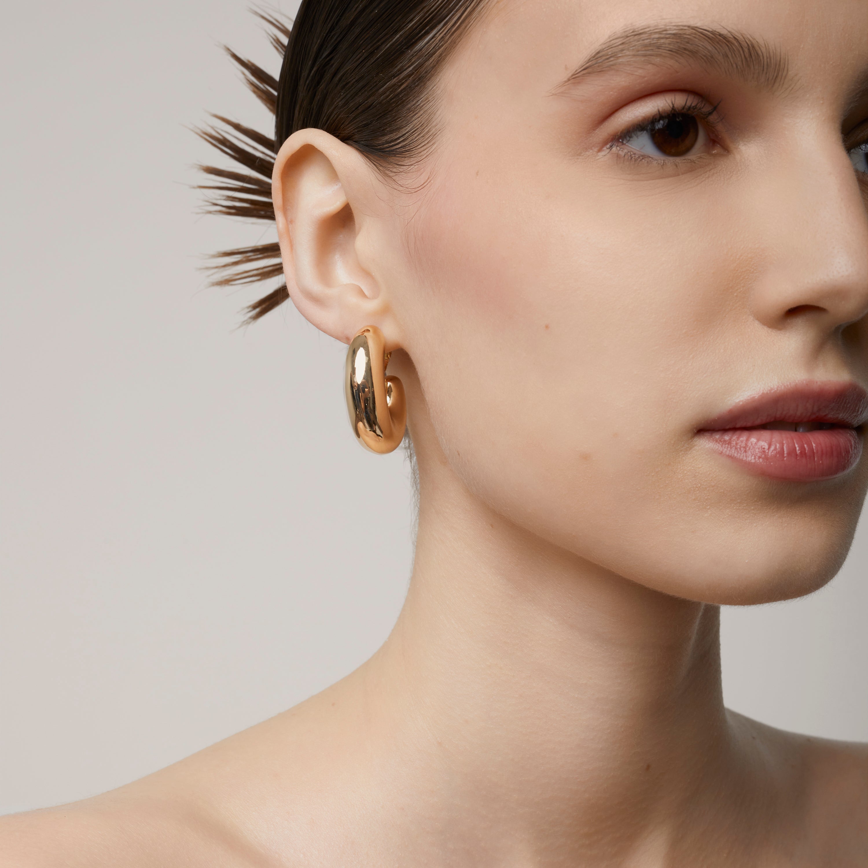 A model wearing the Chunky Oval Hoop Clip On Earrings in Gold - the perfect solution for those with thick, large, sensitive, small, thin, or keloid prone ears. With a convenient screwback clip-on closure, these earrings can be manually adjusted for a secure and comfortable fit that lasts up to 12 hours. This single pair is the perfect addition to any jewelry collection.