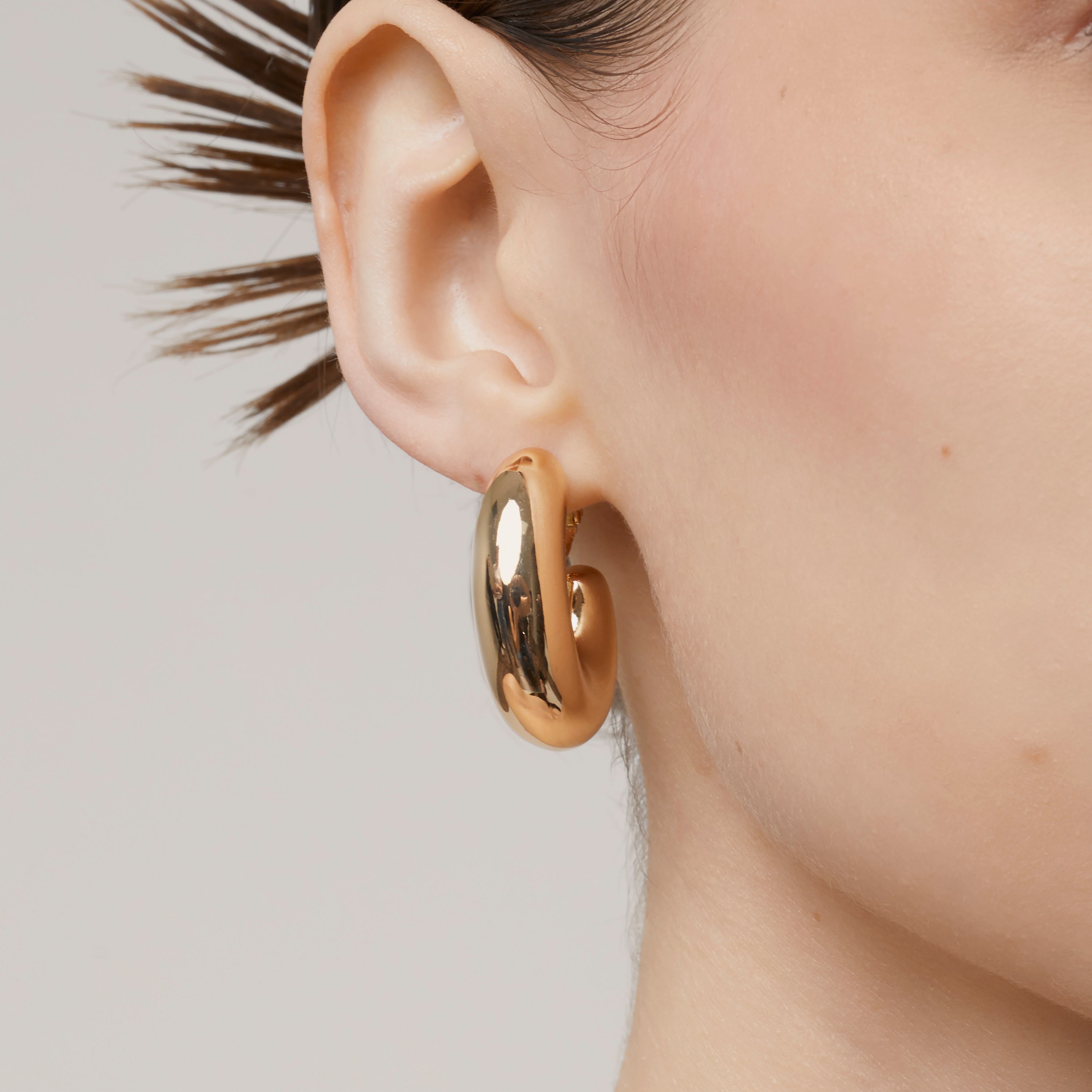 A model wearing the Chunky Oval Hoop Clip On Earrings in Gold - the perfect solution for those with thick, large, sensitive, small, thin, or keloid prone ears. With a convenient screwback clip-on closure, these earrings can be manually adjusted for a secure and comfortable fit that lasts up to 12 hours. This single pair is the perfect addition to any jewelry collection.