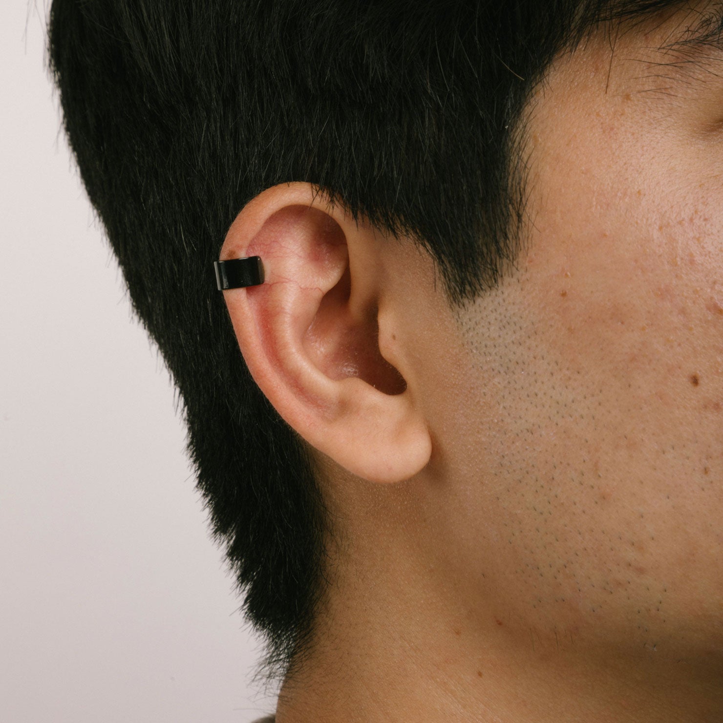 A model wearing the Cigar Band Ear Cuff, available in Black, features a medium-strength secure hold and adjustable closure type. Crafted from Stainless Steel and Titanium, this cuff is suitable for use with all ear types, including thick/large ears, sensitive ears, small/thin ears, and stretched/healing ears. Please note, this item is sold as a single piece and is intended to be worn on the helix of the ear.