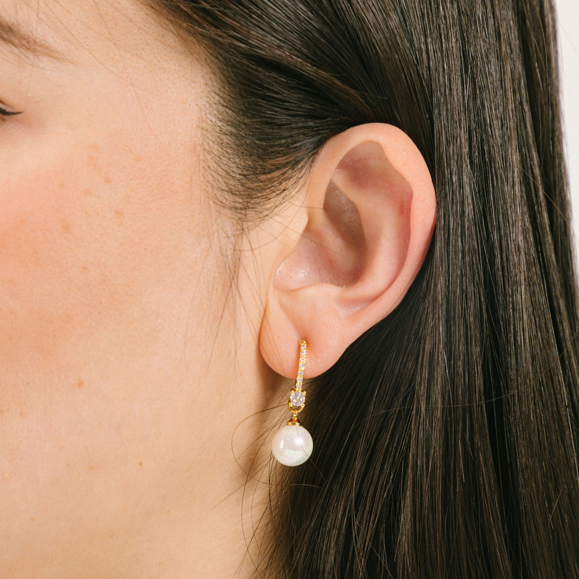 A model wearing the Claire Pearl Clip On Earrings in Gold bear Mosquito Coil Clip-Ons, offering a secure, comfortable fit for all types of ears - from Thick/Large to Sensitive, Small/Thin, and even Stretched/Healing - for up to 24 hours. Meticulously fashioned from 18K Gold plated metal alloy, Cubic Zirconia, and Simulated Faux Pearl, this chic design is Non-Tarnish and Water Resistant.