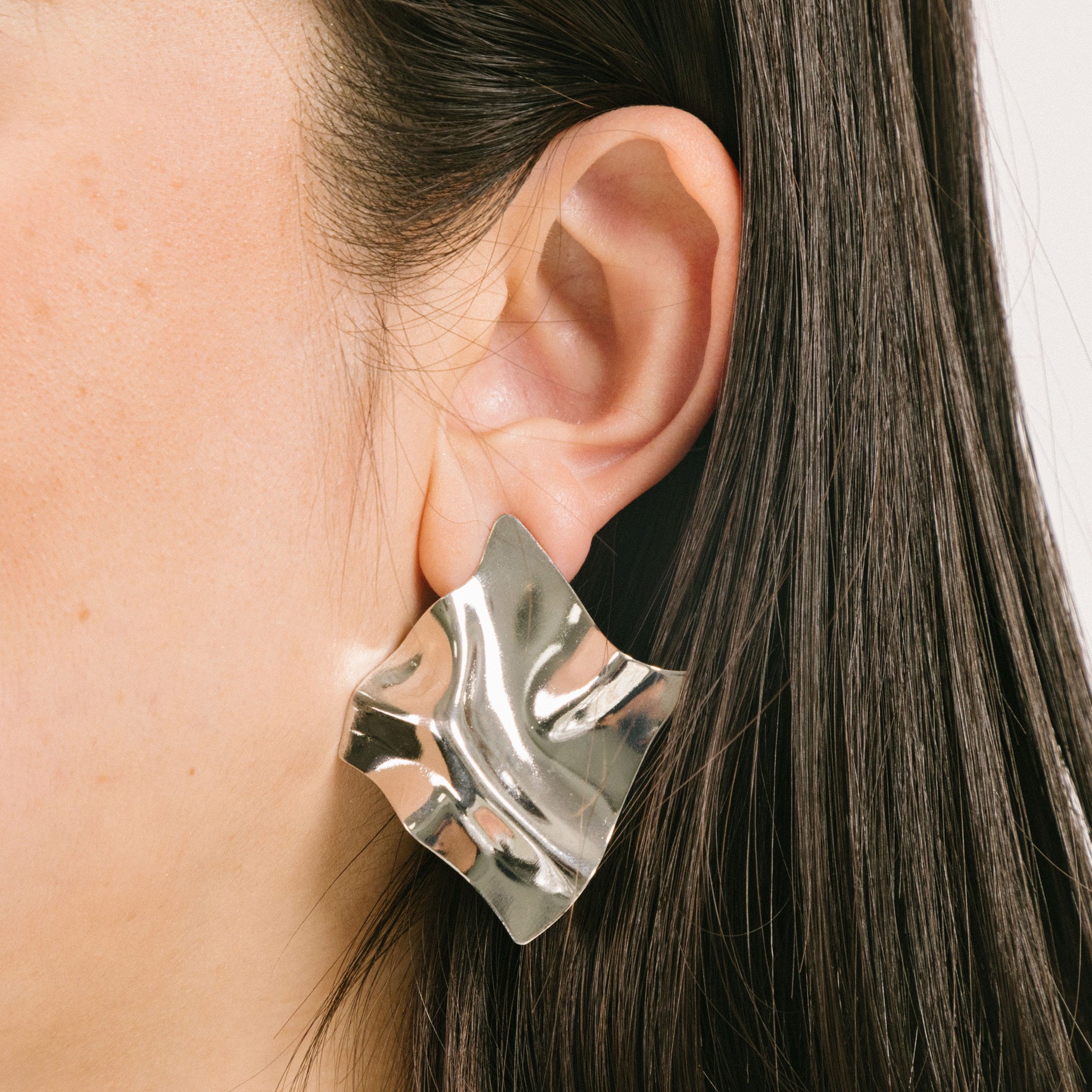 A model wearing the Silver Crinkle Clip On Earrings are designed for all ear types, including those with thick/large, sensitive, small/thin, or stretched/healing ears. This item includes one pair of clip on earrings crafted from silver tone copper alloy. A removable rubber padding ensures secure and comfortable closure.