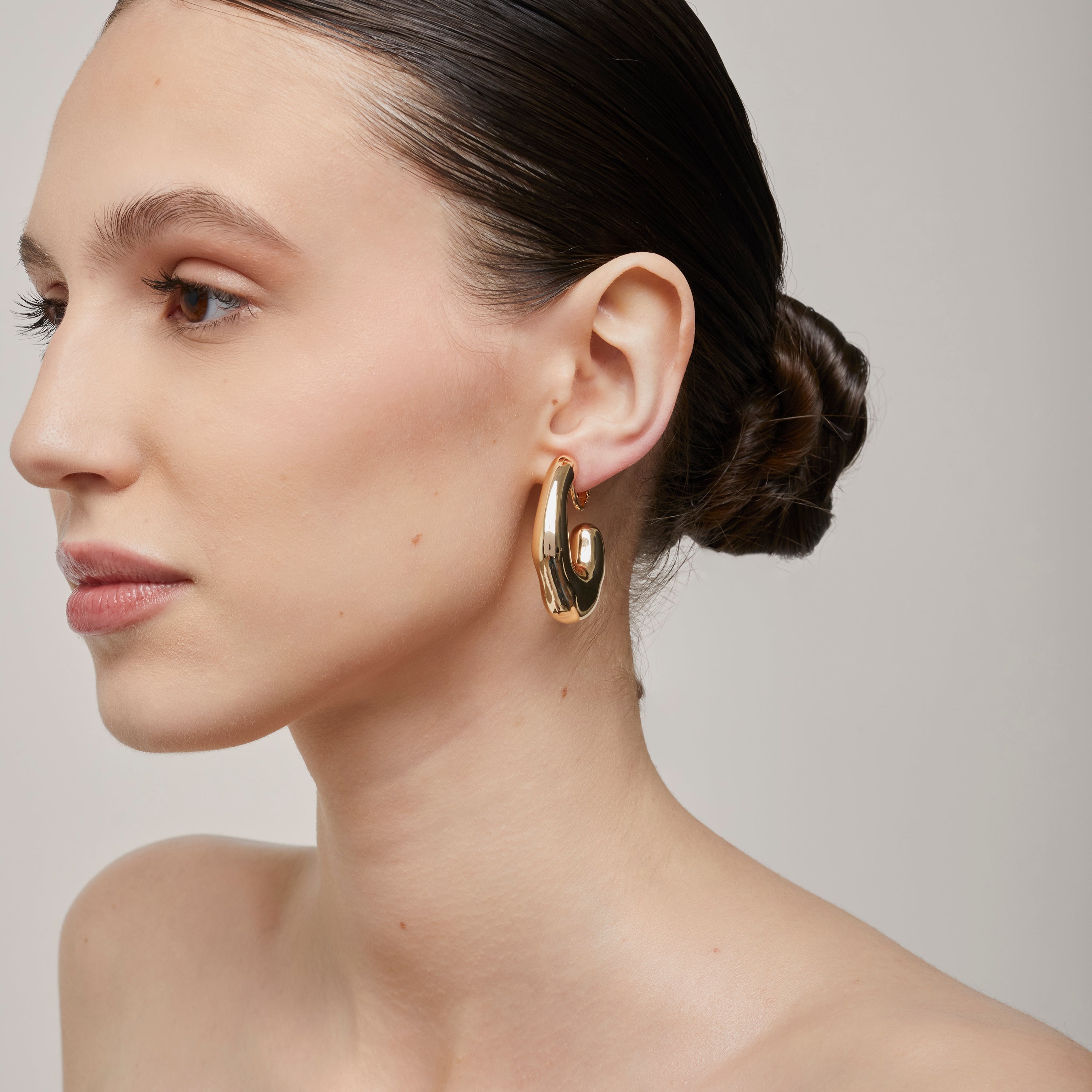 A model wearing the Curvy Hoop Clip On Earrings in Gold. These earrings offer comfort and versatility with a secure screwback closure that suits all ear sizes. Enjoy 8-12 hours of wear without discomfort, suitable for any ear type. Please note that each purchase includes one pair.