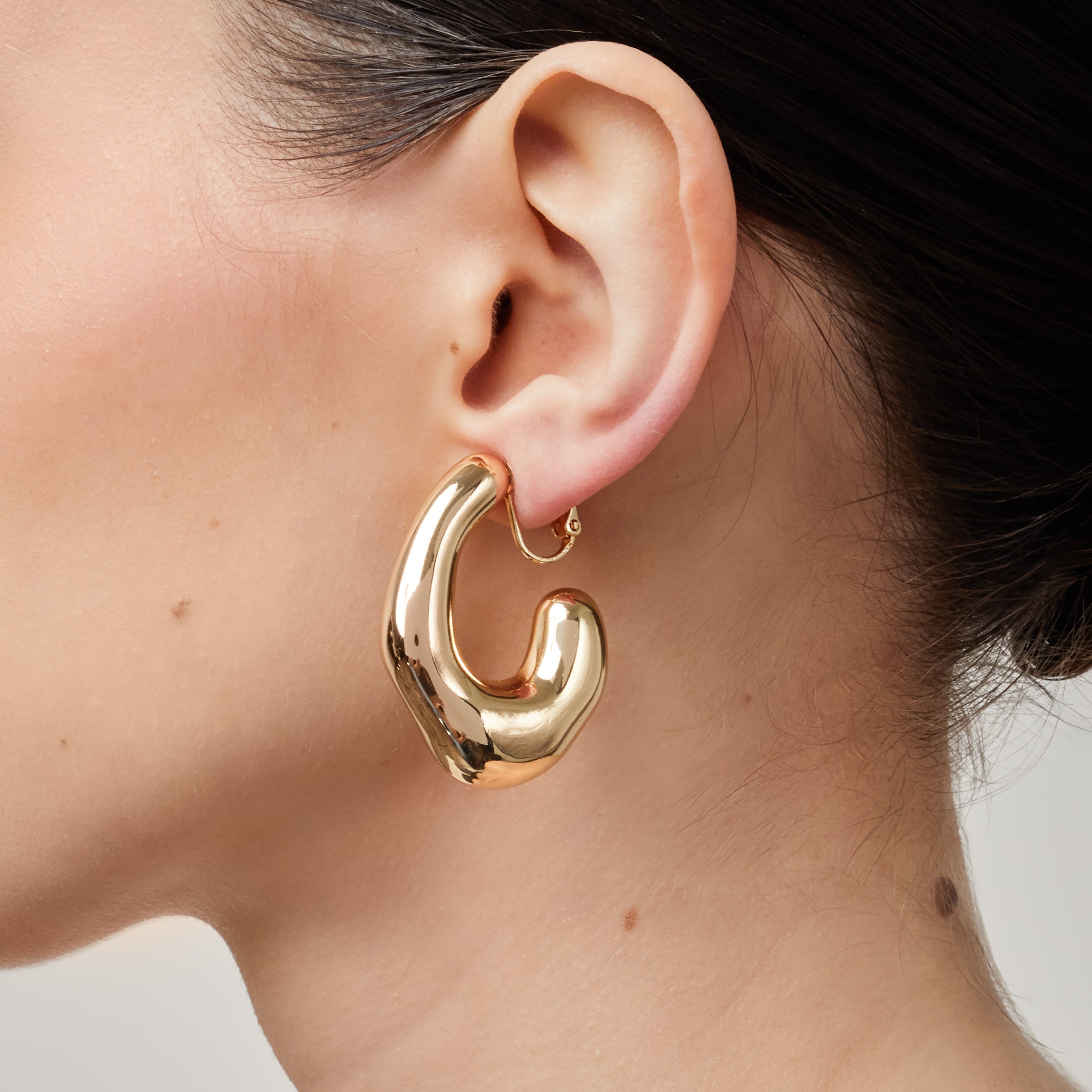 A model wearing the Curvy Hoop Clip On Earrings in Gold. These earrings offer comfort and versatility with a secure screwback closure that suits all ear sizes. Enjoy 8-12 hours of wear without discomfort, suitable for any ear type. Please note that each purchase includes one pair.