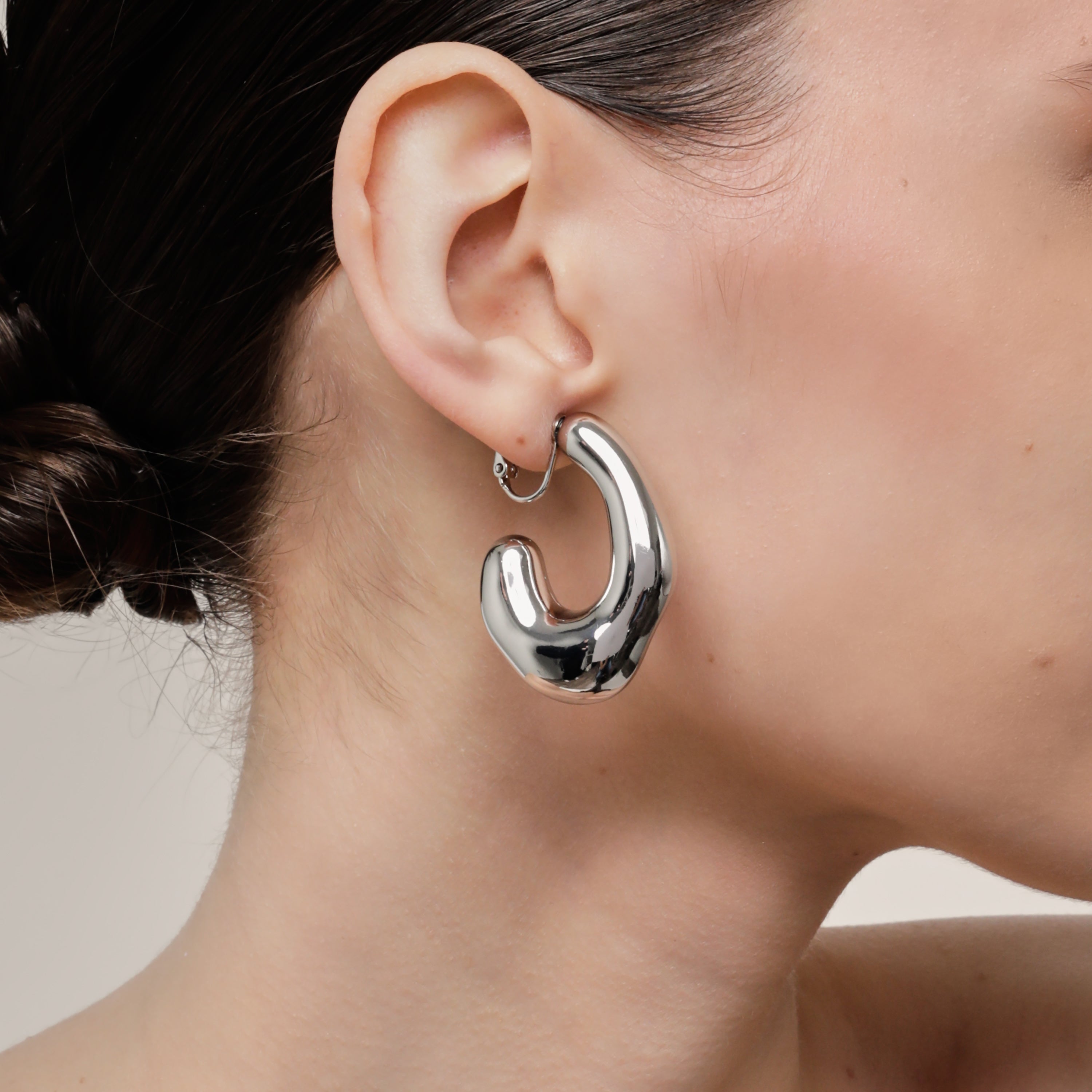 A model wearing the Curvy Hoop Clip On Earrings in Silver. These earrings offer comfort and versatility with a secure screwback closure that suits all ear sizes. Enjoy 8-12 hours of wear without discomfort, suitable for any ear type. Please note that each purchase includes one pair.