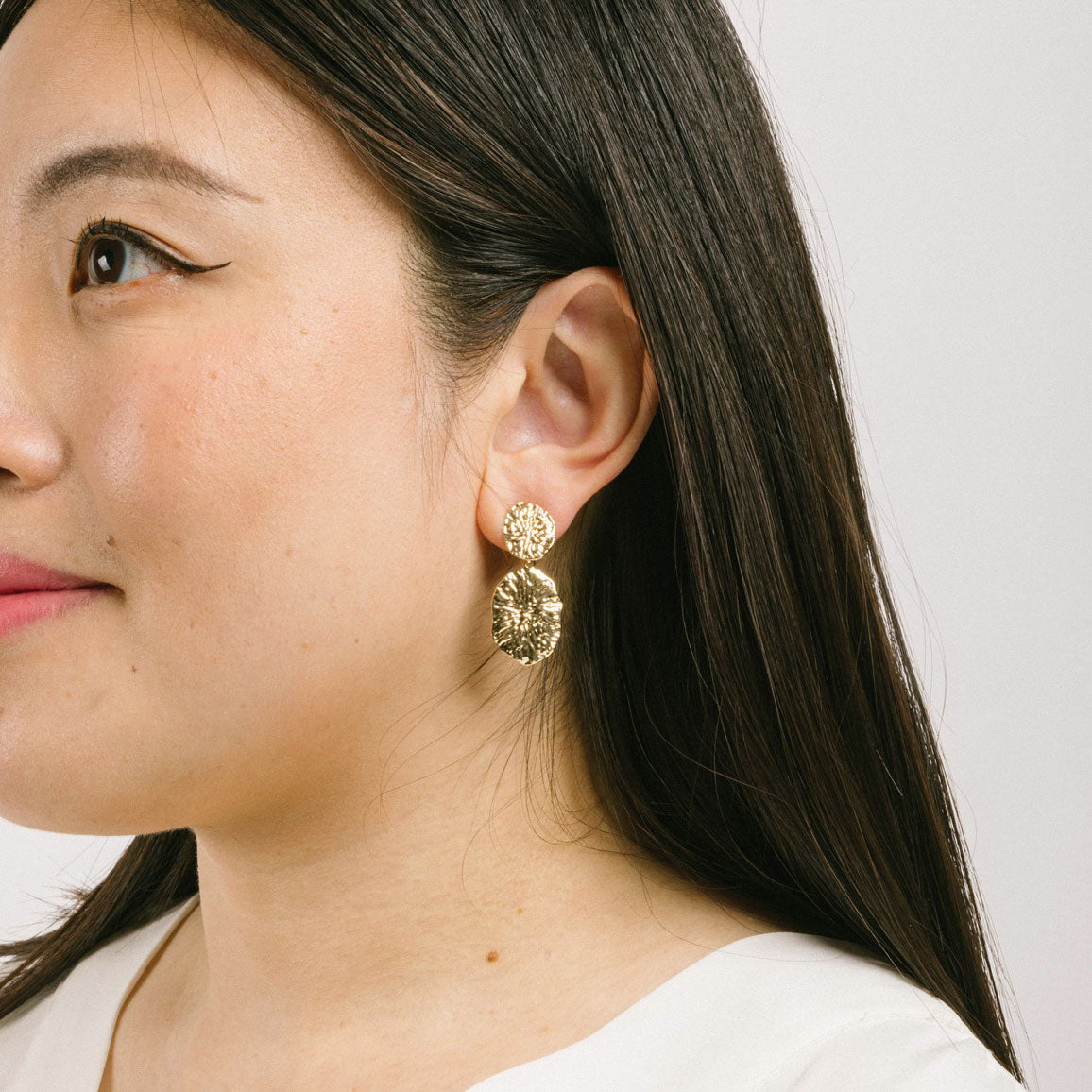 A model wearing the Daiquiri Drop Clip On Earrings in Gold feature a secure, padded clip-on closure ideal for all ear types, offering up to 12 hours of comfortable wear. Crafted from Zinc and Copper alloy, these earrings come as a single pair with removable rubber padding.