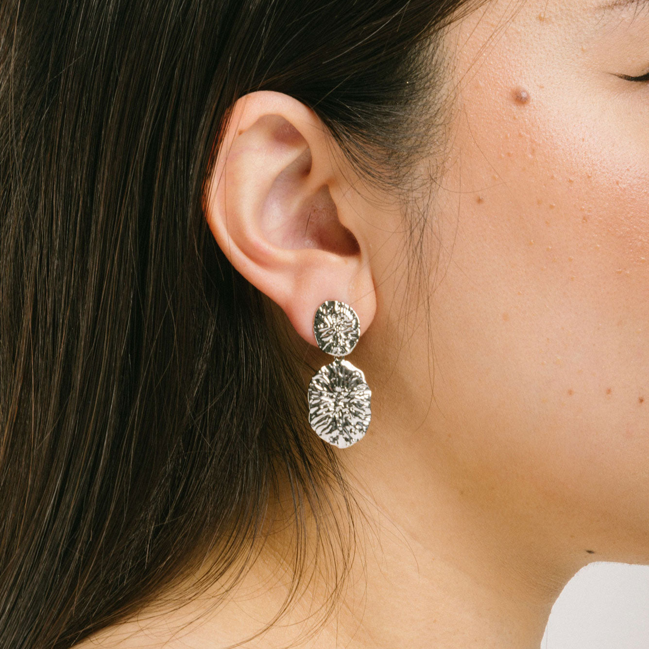 A model wearing the Daiquiri Drop Clip On Earrings in Silver feature a secure, padded clip-on closure ideal for all ear types, offering up to 12 hours of comfortable wear. Crafted from Zinc and Copper alloy, these earrings come as a single pair with removable rubber padding.