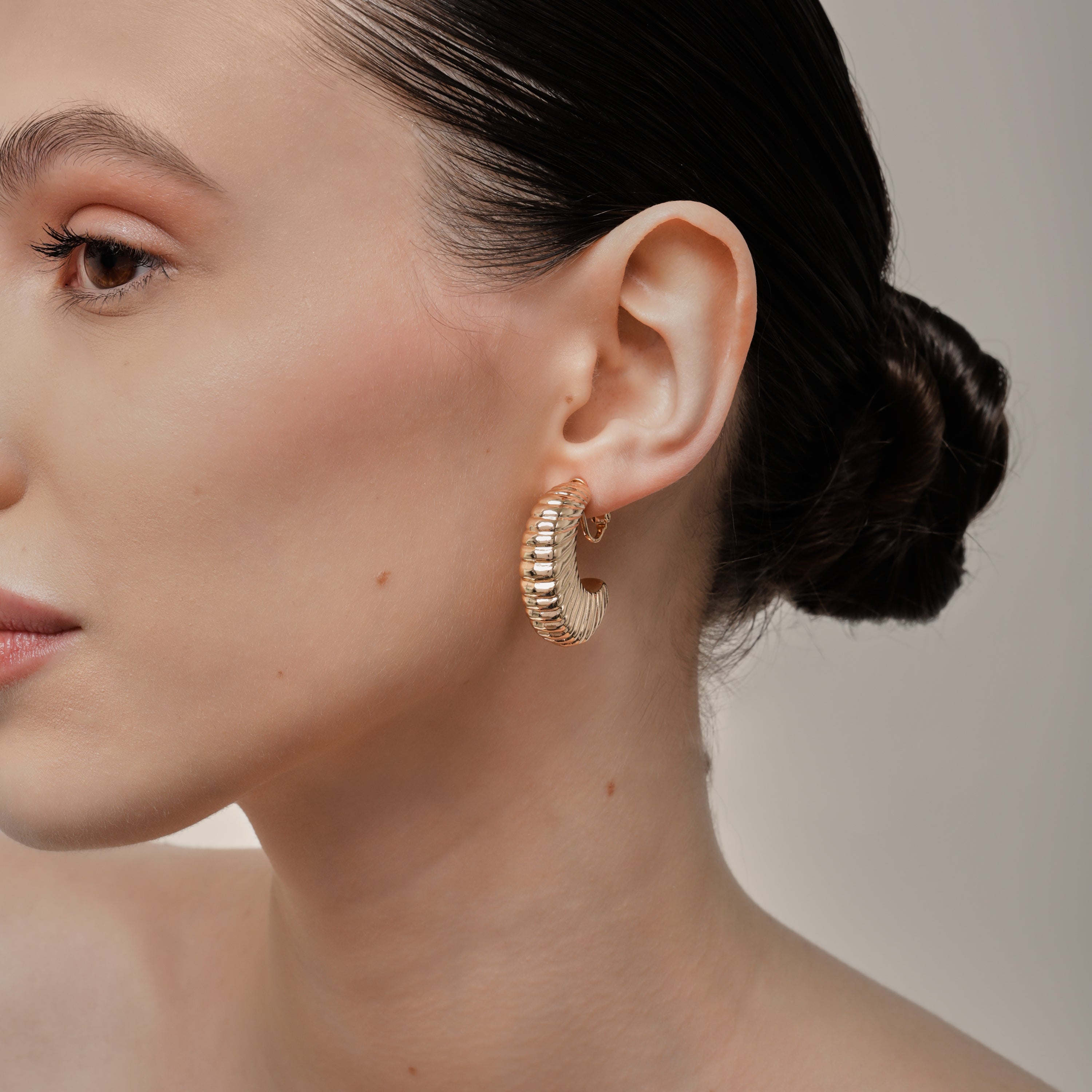 A model wearing the Deco Hoop Clip On Earrings in Gold. Designed with a screwback closure, these earrings provide a secure hold for all types of ears. Whether you have thick, sensitive, small, or keloid prone ears, these earrings can be manually adjusted to fit your size. Enjoy 8-12 hours of wear in these elegant, versatile earrings.