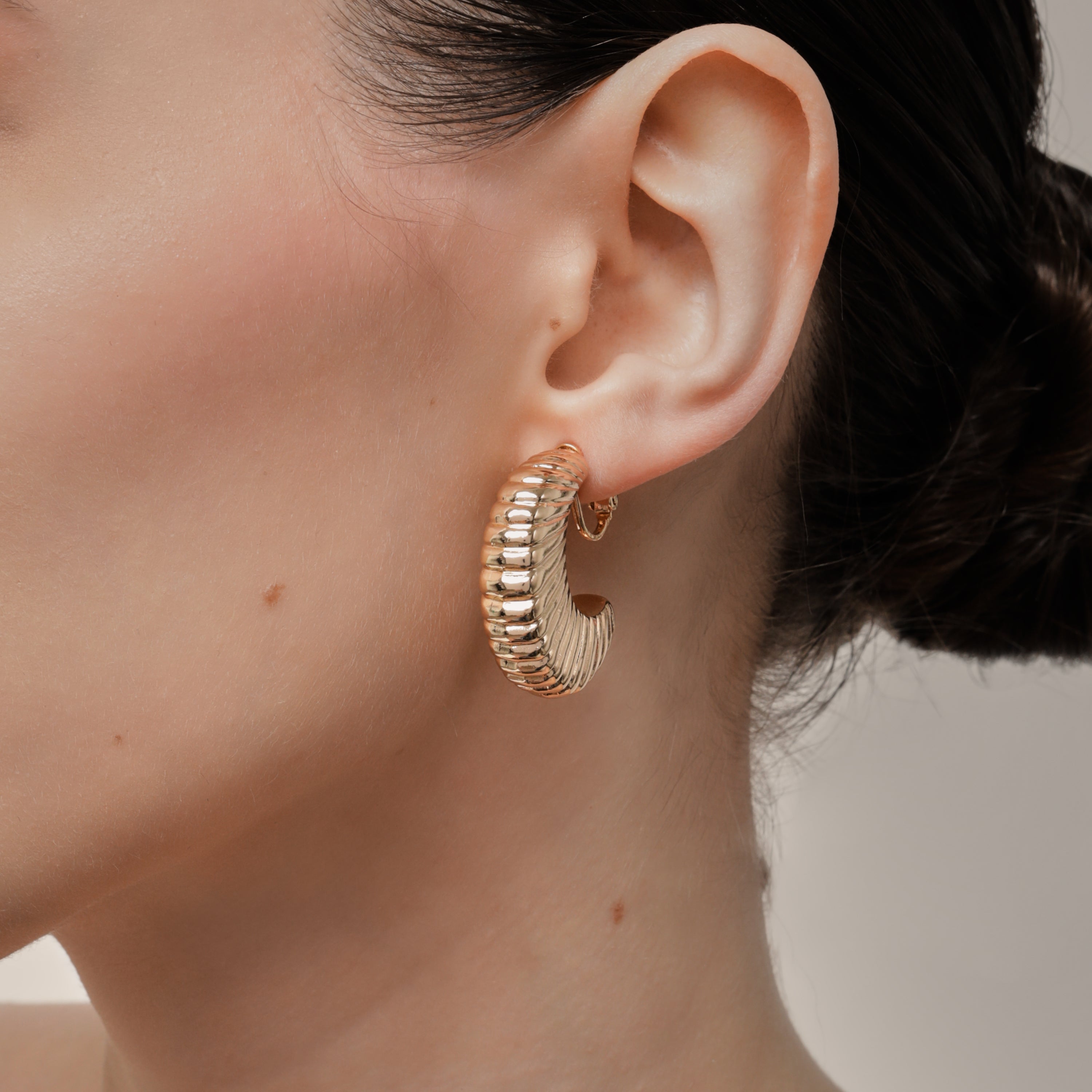 A model wearing the Deco Hoop Clip On Earrings in Gold. Designed with a screwback closure, these earrings provide a secure hold for all types of ears. Whether you have thick, sensitive, small, or keloid prone ears, these earrings can be manually adjusted to fit your size. Enjoy 8-12 hours of wear in these elegant, versatile earrings.