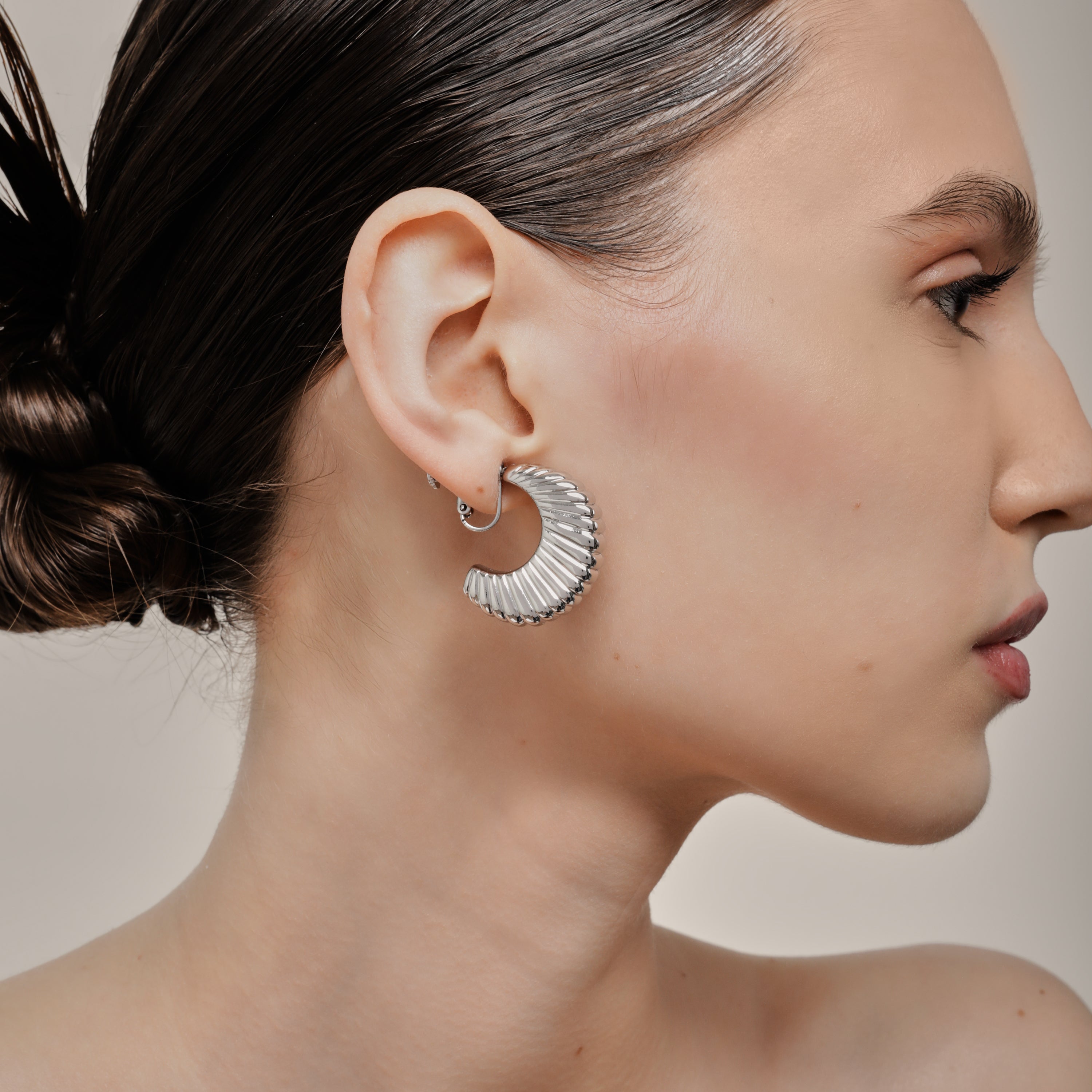 A model wearing the Deco Hoop Clip On Earrings in Silver. Designed with a screwback closure, these earrings provide a secure hold for all types of ears. Whether you have thick, sensitive, small, or keloid prone ears, these earrings can be manually adjusted to fit your size. Enjoy 8-12 hours of wear in these elegant, versatile earrings.
