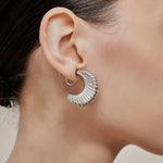 A model wearing the Deco Hoop Clip On Earrings in Silver. Designed with a screwback closure, these earrings provide a secure hold for all types of ears. Whether you have thick, sensitive, small, or keloid prone ears, these earrings can be manually adjusted to fit your size. Enjoy 8-12 hours of wear in these elegant, versatile earrings.
