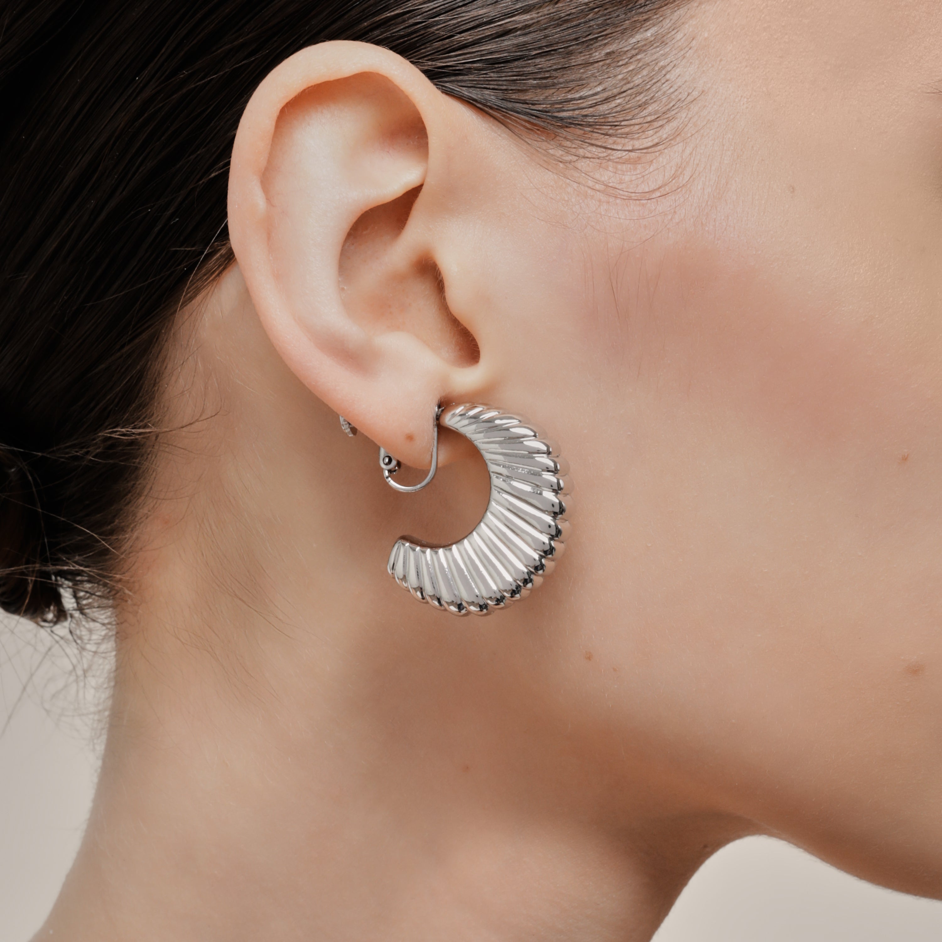 A model wearing the Deco Hoop Clip On Earrings in Silver. Designed with a screwback closure, these earrings provide a secure hold for all types of ears. Whether you have thick, sensitive, small, or keloid prone ears, these earrings can be manually adjusted to fit your size. Enjoy 8-12 hours of wear in these elegant, versatile earrings.