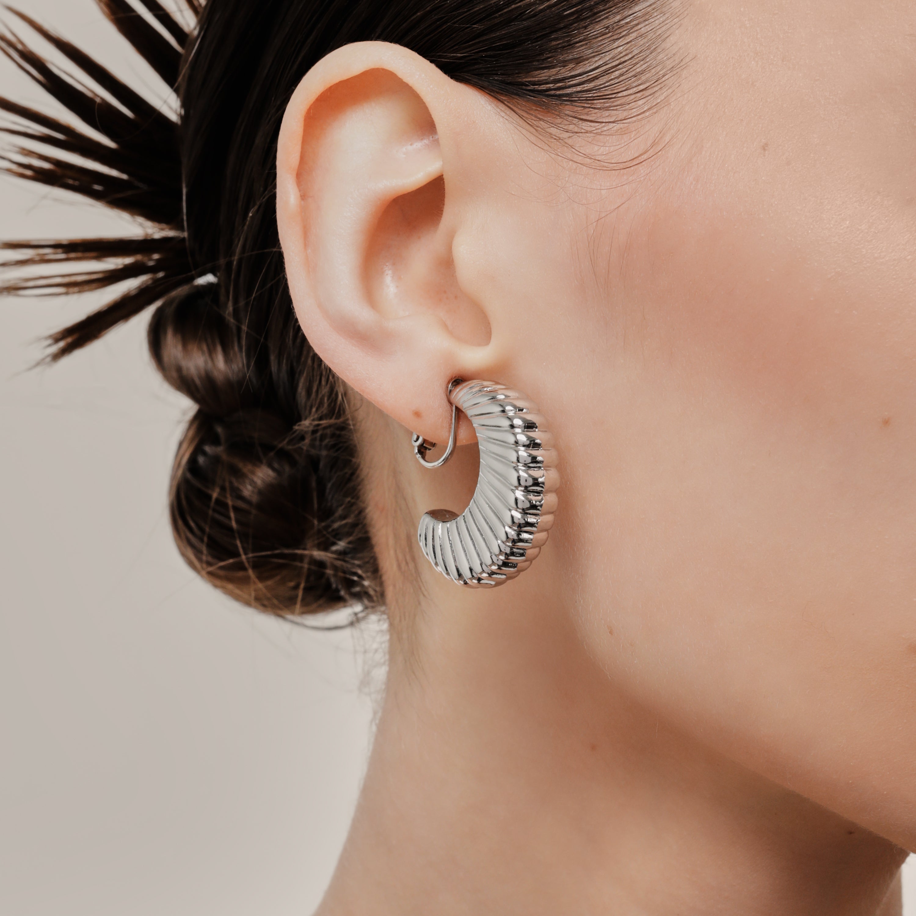 A model wearing the Deco Hoop Clip On Earrings in Silver. Designed with a screwback closure, these earrings provide a secure hold for all types of ears. Whether you have thick, sensitive, small, or keloid prone ears, these earrings can be manually adjusted to fit your size. Enjoy 8-12 hours of wear in these elegant, versatile earrings.
