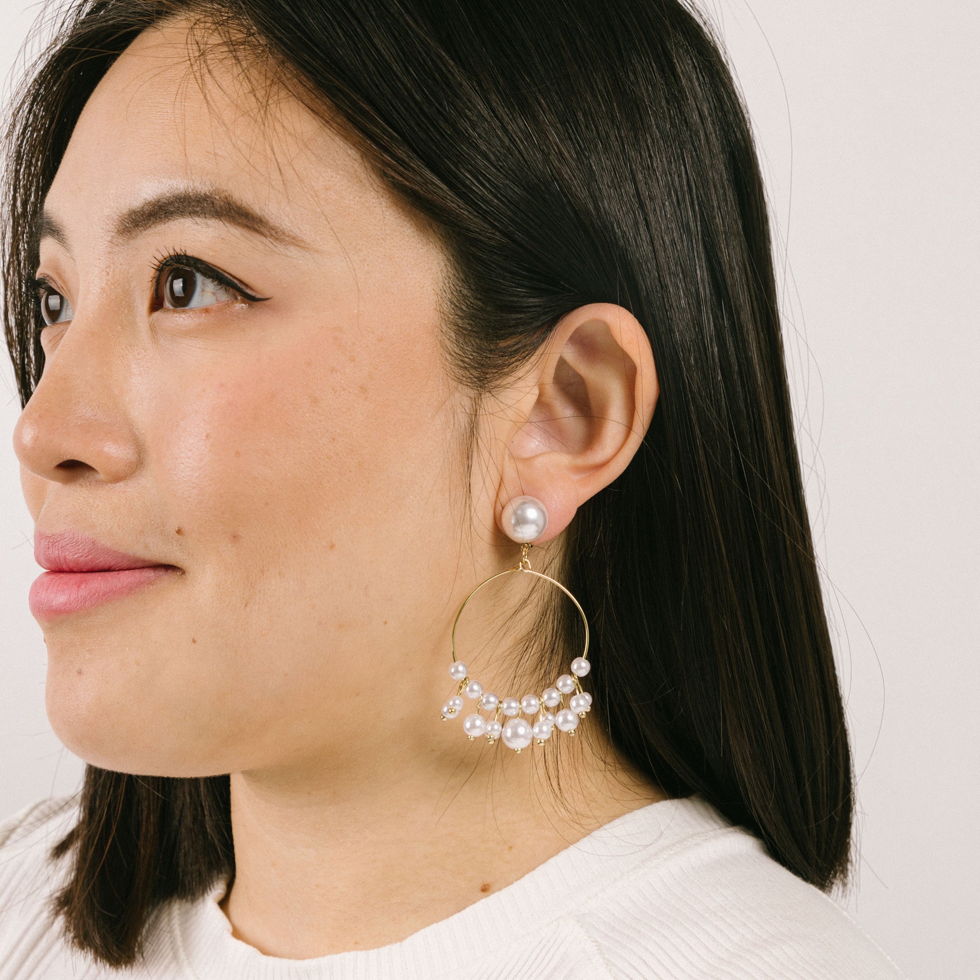 A model wearing the Delphine Clip On Earrings feature a gold-tone copper alloy and a Simulated Faux Pearl. The closure type is a padded clip-on earring with removable rubber padding, making it ideal for all ear types (including thick/large ears, sensitive ears, small/thin ears, and stretched/healing ears). You can expect a secure hold for up to 12 hours of comfortable wear. Please note that this item is sold as a single pair.