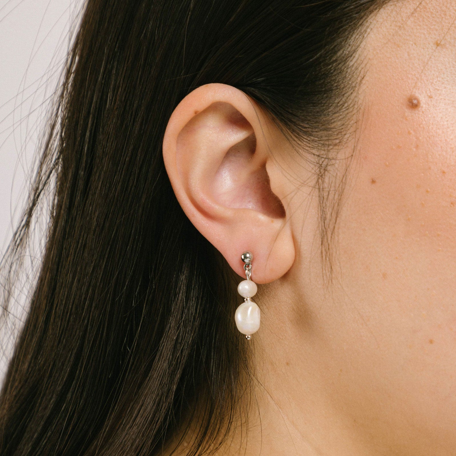 A model wearing the Silver Plated Brass Duo Pearl Clip-On Earrings features a mosquito coil clip-on design, making it ideal for all ear types including those with thick/large, sensitive, small/thin, and stretched/healing ears. These earrings comfortably allow for up to 24 hours of wear and have a medium secure hold with adjustable padding. The freshwater pearls used in these earrings may vary slightly in size and colour.