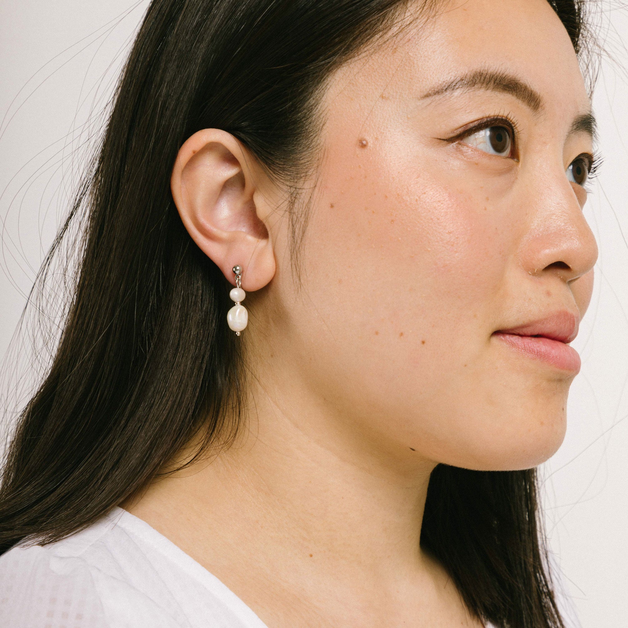 A model wearing the Silver Plated Brass Duo Pearl Clip-On Earrings features a mosquito coil clip-on design, making it ideal for all ear types including those with thick/large, sensitive, small/thin, and stretched/healing ears. These earrings comfortably allow for up to 24 hours of wear and have a medium secure hold with adjustable padding. The freshwater pearls used in these earrings may vary slightly in size and colour.