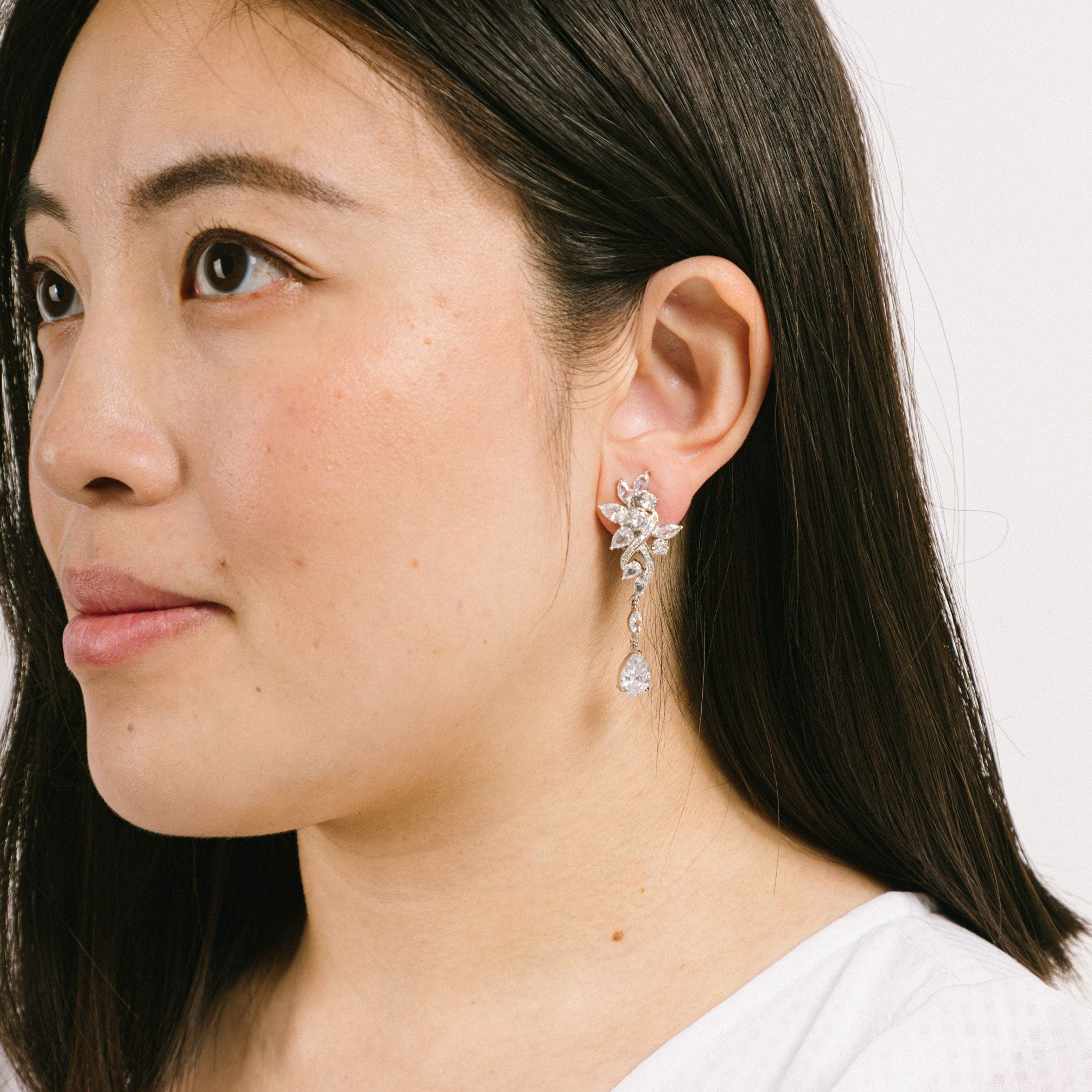 A model wearing the Evelyn Clip-On Earrings offer a secure fit for all ear types, ranging from thick/large ears to small/thin ears. With an average comfortable wear duration of 8-12 hours, these earrings have silver plated copper alloy components and feature a cubic zirconia setting. Additionally, the rubber padding is removable. Please note that this item is one pair.