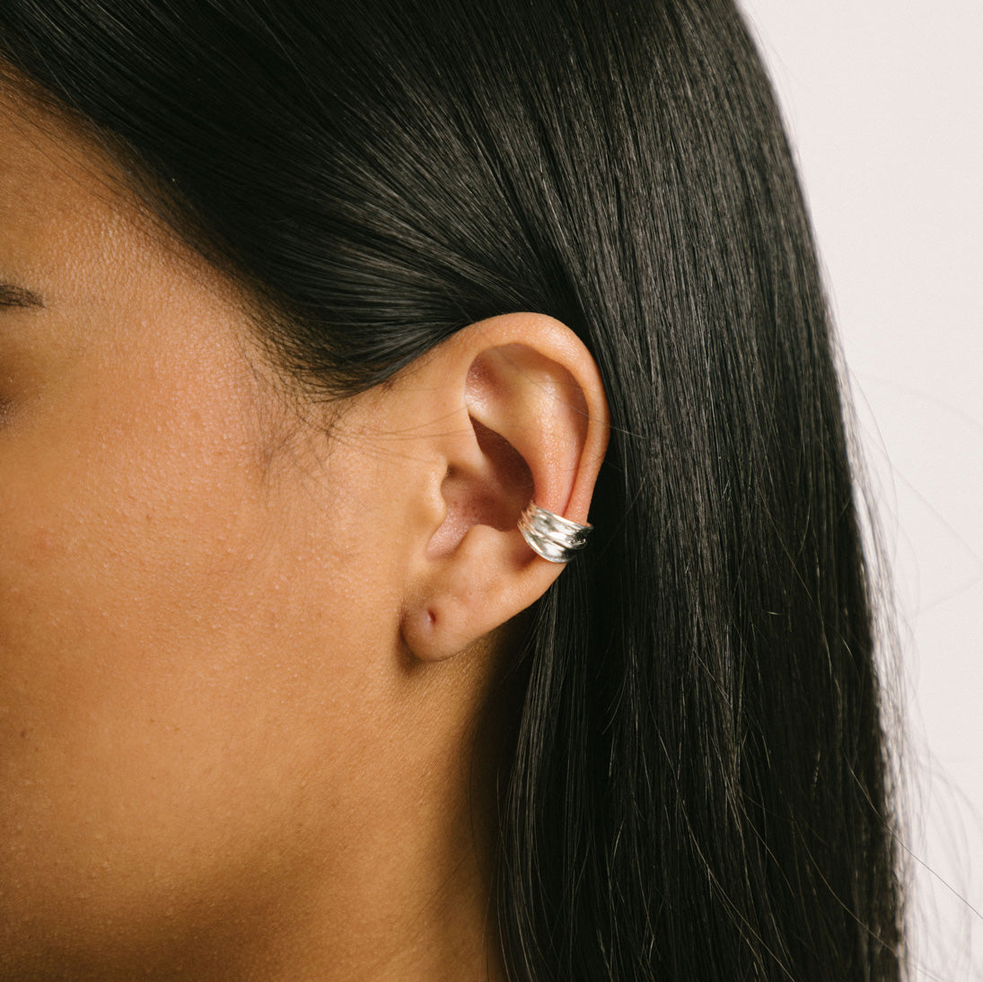 A model wearing the Mona Ear Cuff in Silver is designed for all ear types, providing a comfortable, medium secure hold for up to 24 hours. It features the ability to adjust with gentle squeezing, crafted with 925 Sterling Silver in one piece.