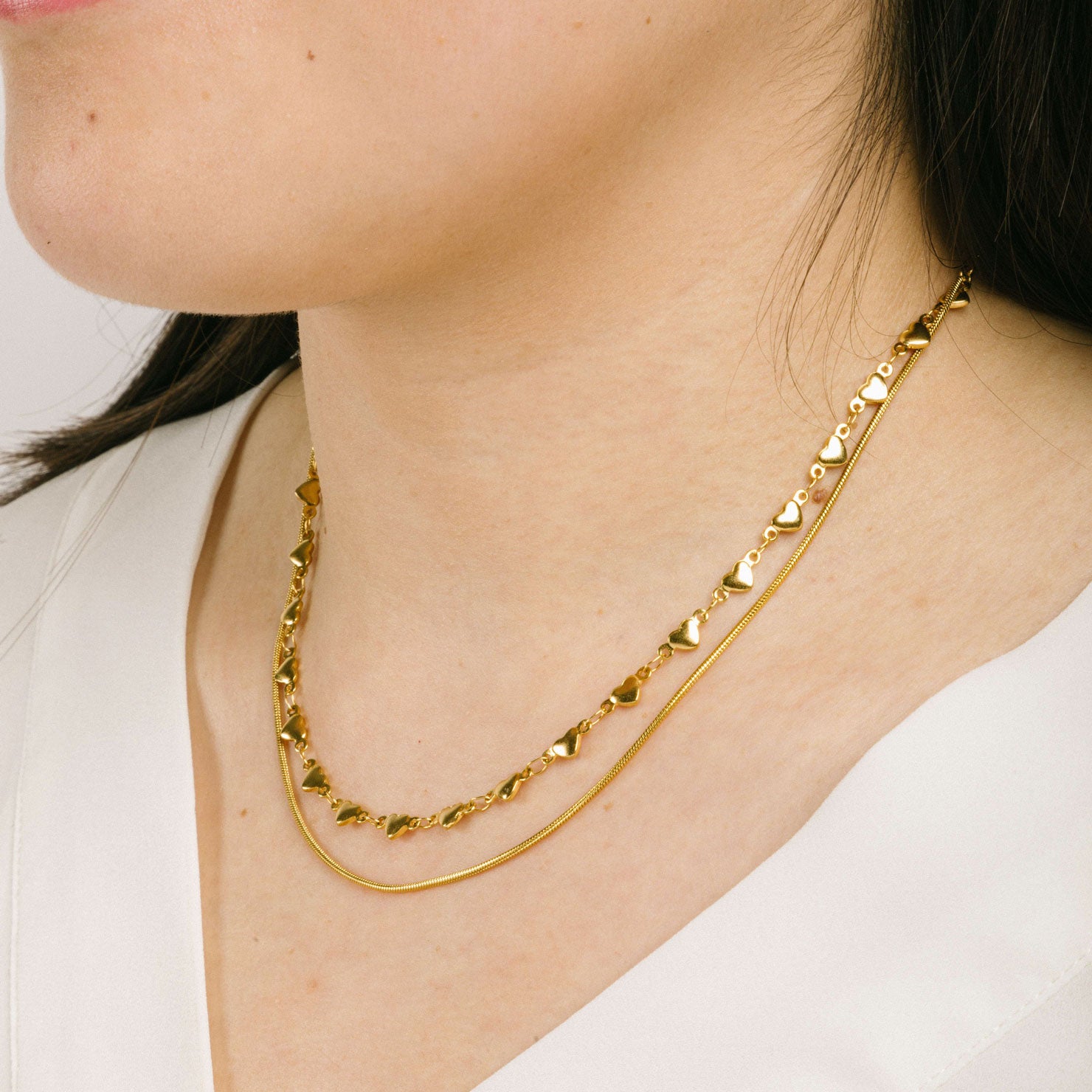 A model wearing the Heart Chain Double Layered Necklace is crafted from 14K Gold Plated Stainless Steel, which is water resistant and non-tarnishing. The necklace can be adjusted for a perfect fit. Please Note: This product is one necklace.