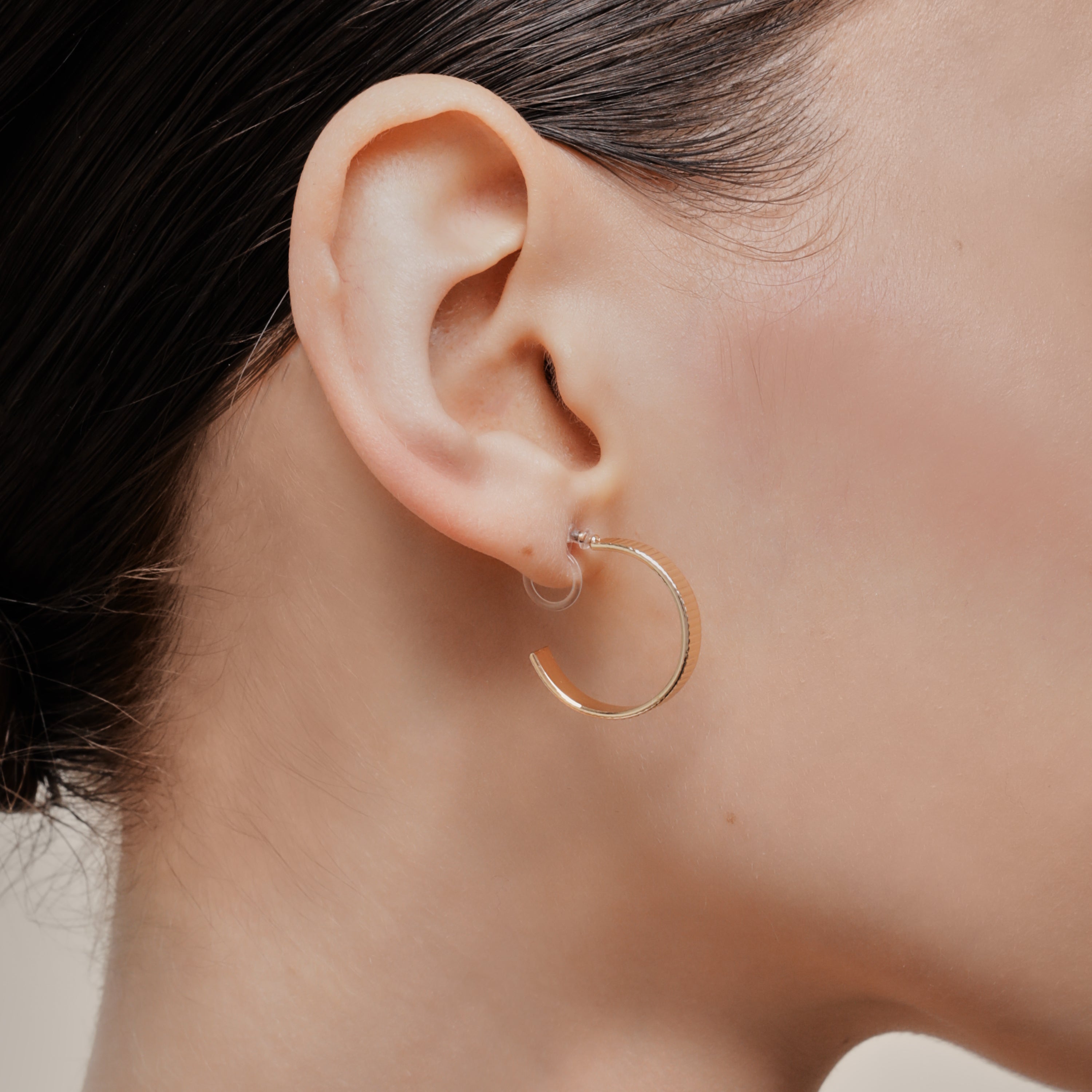A model wearing the Pleated Hoop Clip On Earrings in Gold offer a resin clip-on closure for a secure and comfortable fit for a variety of ear sizes and sensitivities. With an average wear duration of 8-12 hours, these earrings are perfect for all-day wear. Please note that this listing is for a single pair
