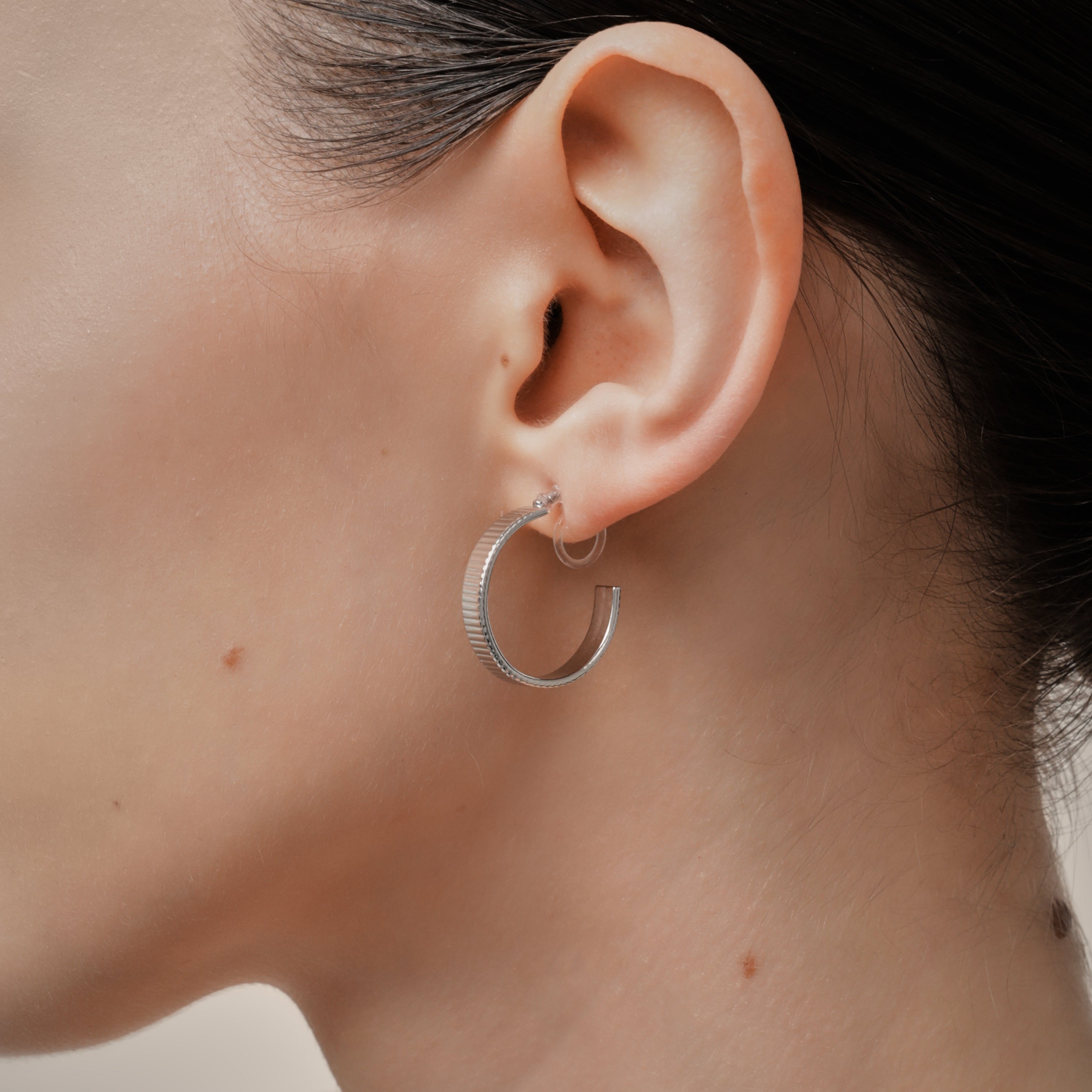 A model wearing the Pleated Hoop Clip On Earrings in Silver offer a resin clip-on closure for a secure and comfortable fit for a variety of ear sizes and sensitivities. With an average wear duration of 8-12 hours, these earrings are perfect for all-day wear. Please note that this listing is for a single pair