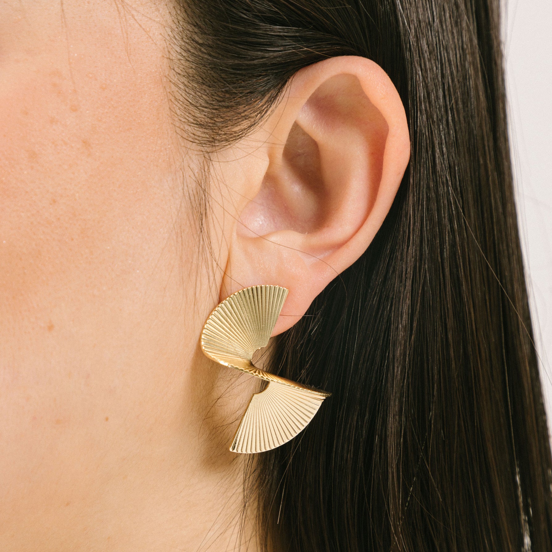 A model wearing the Ribbed Swirl Clip-On Earrings are crafted from a gold-tone copper alloy. These earrings provide a secure hold for up to 12 hours of wear and can comfortably accommodate all ear types, including those with thick or sensitive ears. The clip-on design is complete with removable rubber padding, offering a customized fit with no adjustments necessary. Please note- this item contains one pair.