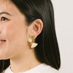 A model wearing the Ribbed Swirl Clip-On Earrings are crafted from a gold-tone copper alloy. These earrings provide a secure hold for up to 12 hours of wear and can comfortably accommodate all ear types, including those with thick or sensitive ears. The clip-on design is complete with removable rubber padding, offering a customized fit with no adjustments necessary. Please note- this item contains one pair.