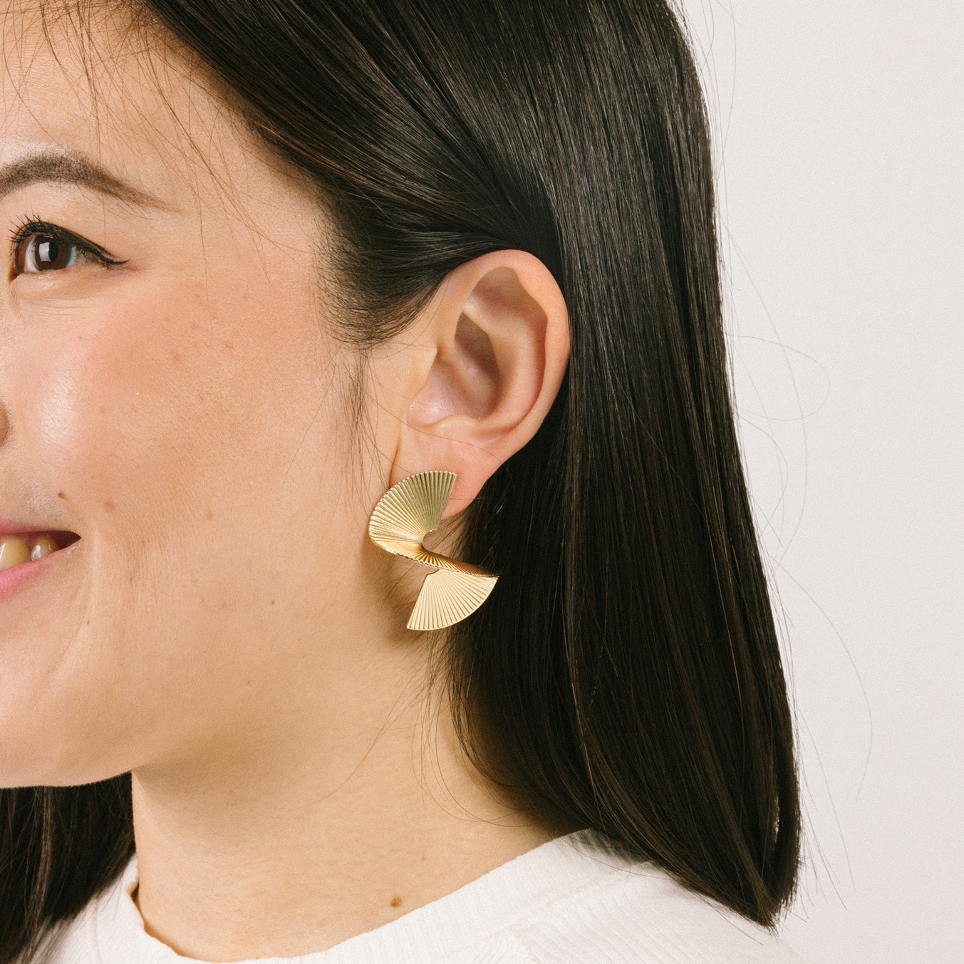 A model wearing the Ribbed Swirl Clip-On Earrings are crafted from a gold-tone copper alloy. These earrings provide a secure hold for up to 12 hours of wear and can comfortably accommodate all ear types, including those with thick or sensitive ears. The clip-on design is complete with removable rubber padding, offering a customized fit with no adjustments necessary. Please note- this item contains one pair.