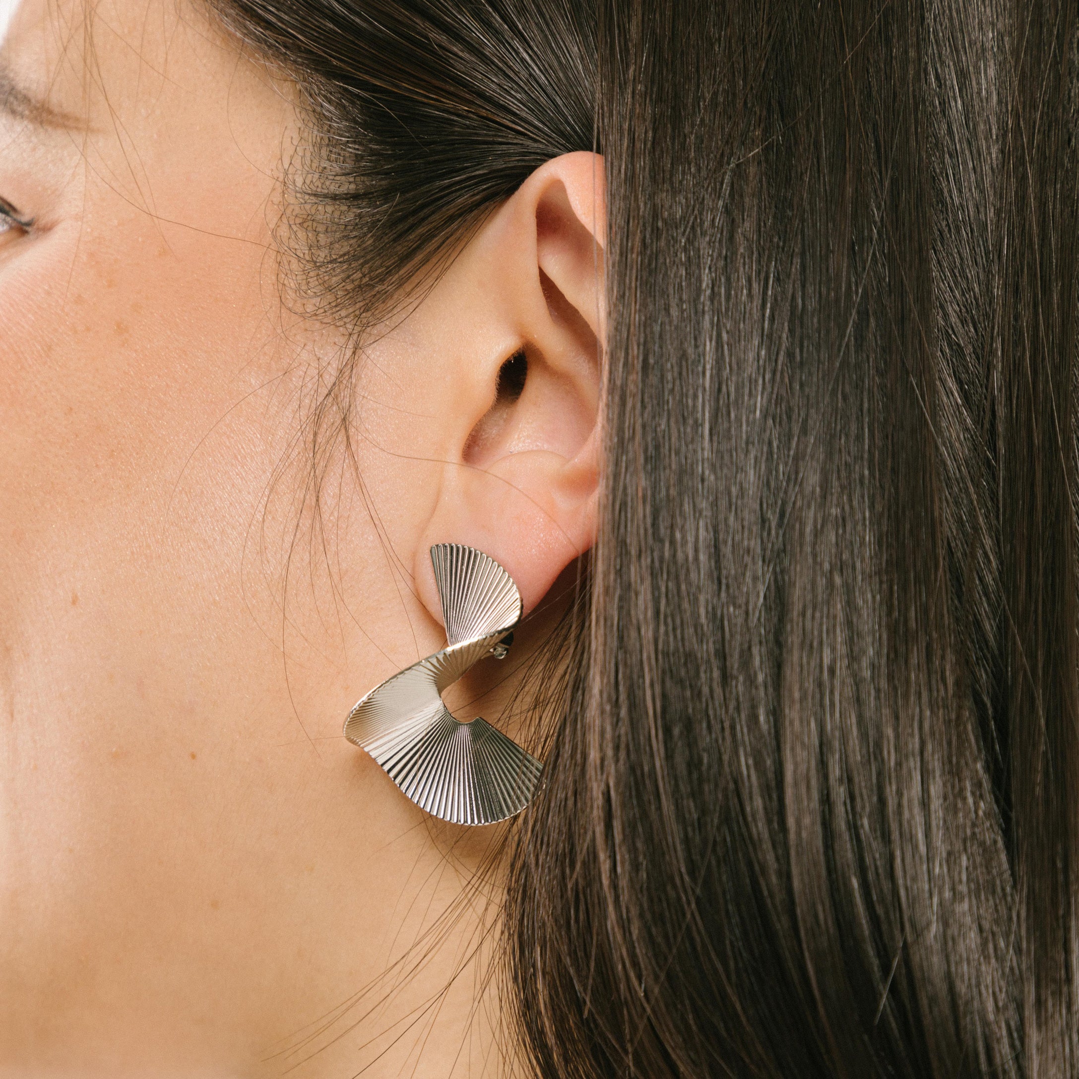 A model wearing the Ribbed Swirl Clip On Earrings in Silver feature a padded clip-on closure that can be worn comfortably for 8-12 hours. Made with silver tone copper alloy, these earrings provide a secure hold that can be adjusted to fit all ear types, such as thick/large, sensitive, small/thin, and stretched/healing. Please note, item is sold as one pair.