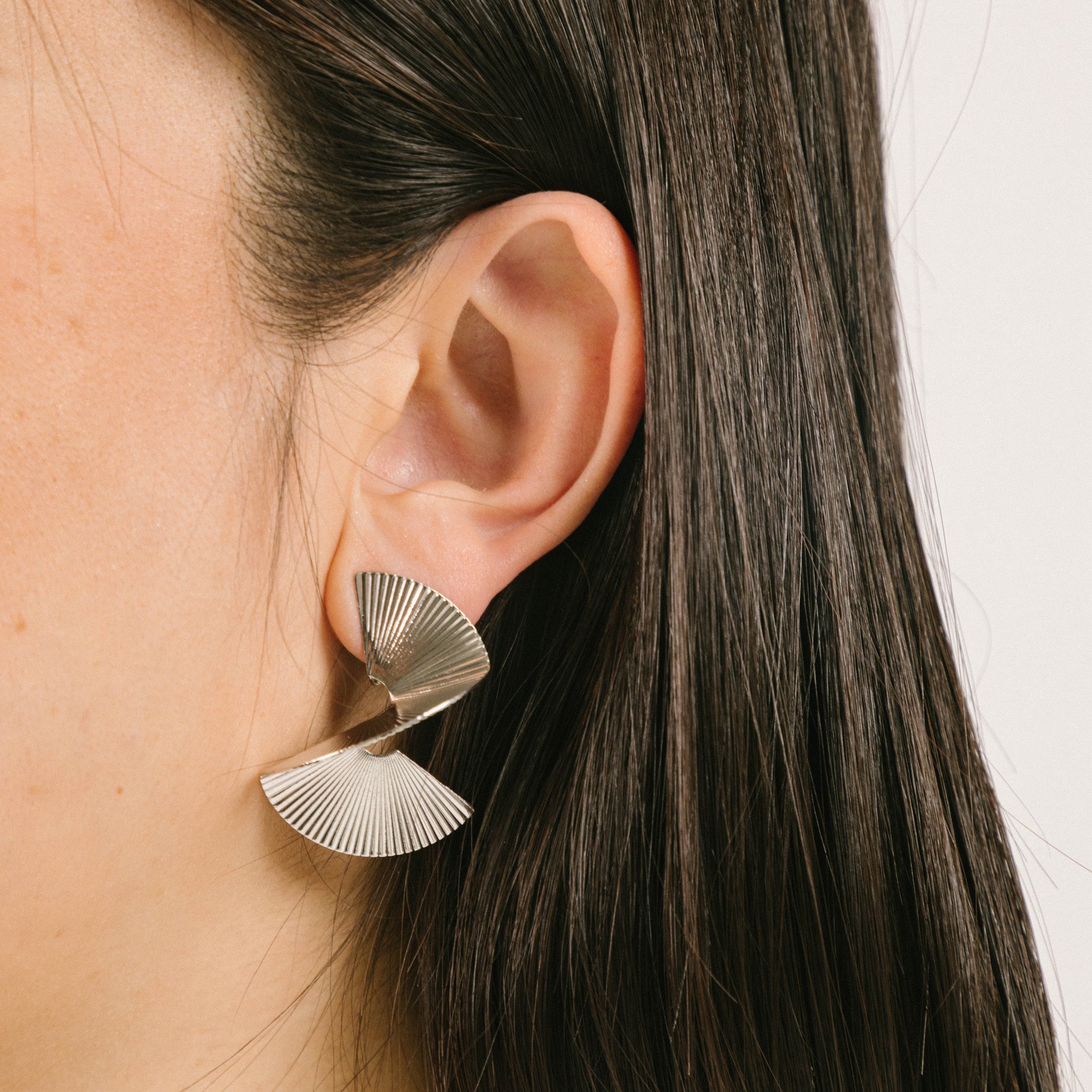 A model wearing the Ribbed Swirl Clip On Earrings in Silver feature a padded clip-on closure that can be worn comfortably for 8-12 hours. Made with silver tone copper alloy, these earrings provide a secure hold that can be adjusted to fit all ear types, such as thick/large, sensitive, small/thin, and stretched/healing. Please note, item is sold as one pair.