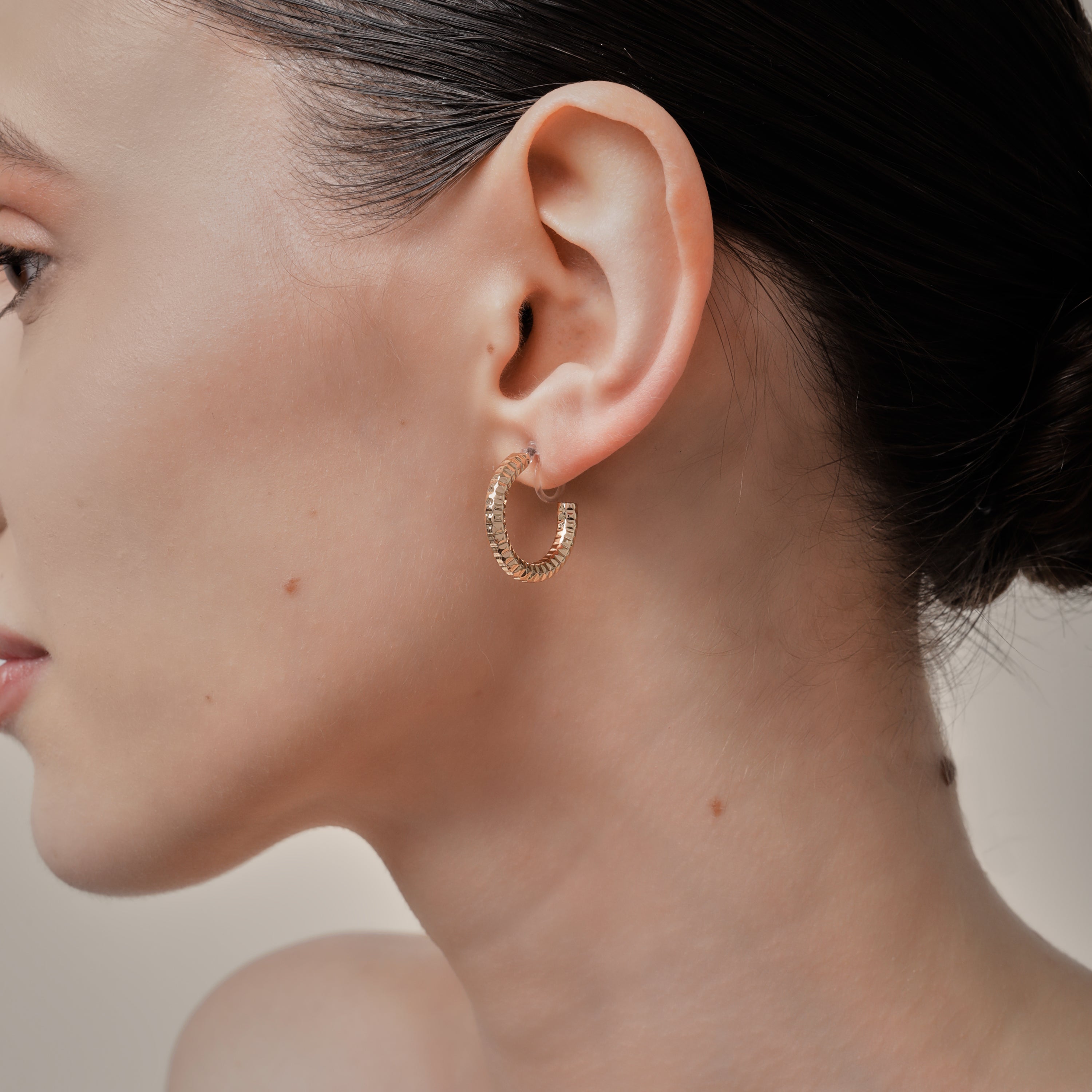 A model wearing the Ribbed Hoop Clip On Earrings in Gold. Made with a Resin Clip-On closure, these earrings provide a secure hold for 8-12 hours and are suitable for all ear sizes and types. Elevate your look with these versatile and long-lasting earrings. Set includes one pair.