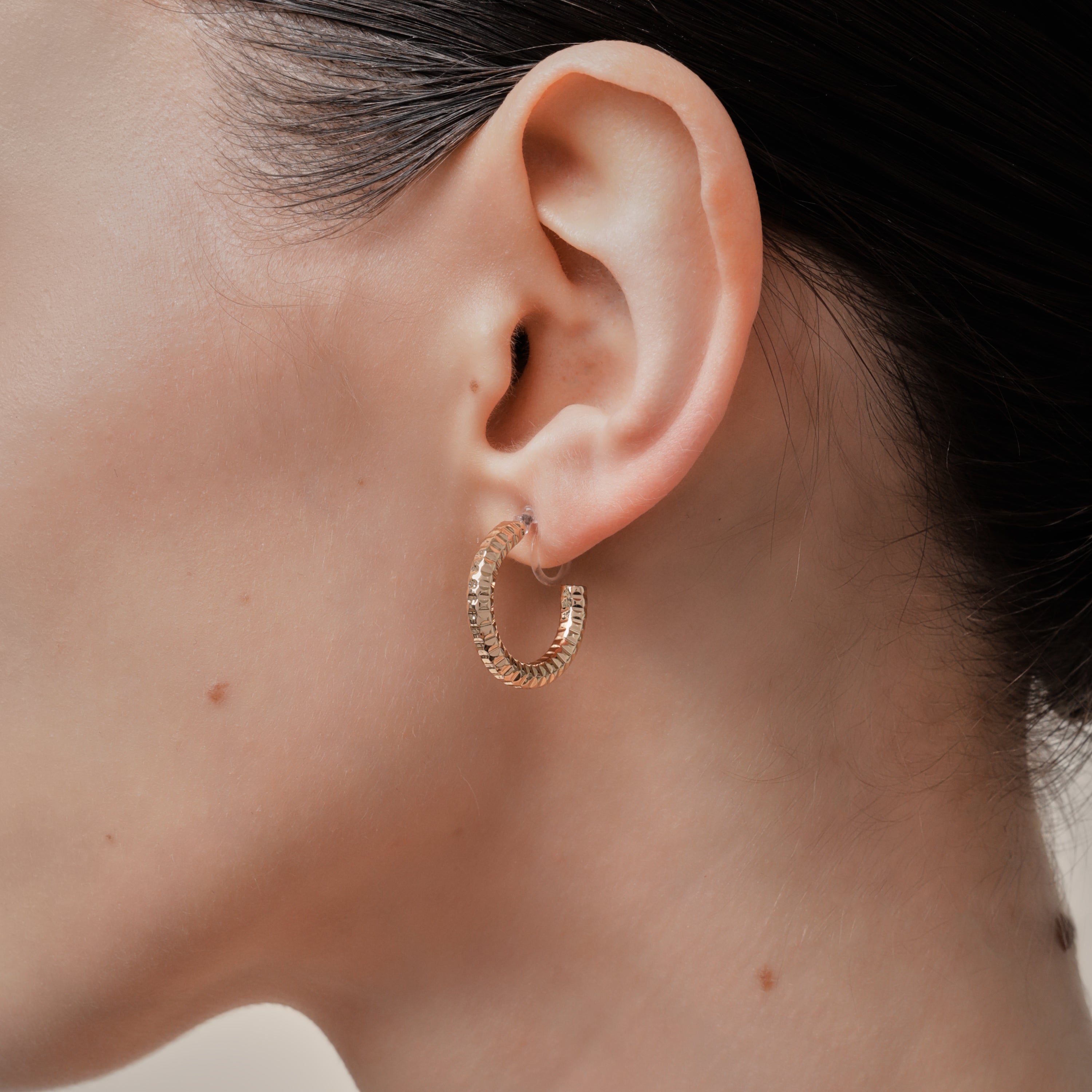 A model wearing the Ribbed Hoop Clip On Earrings in Gold. Made with a Resin Clip-On closure, these earrings provide a secure hold for 8-12 hours and are suitable for all ear sizes and types. Elevate your look with these versatile and long-lasting earrings. Set includes one pair.