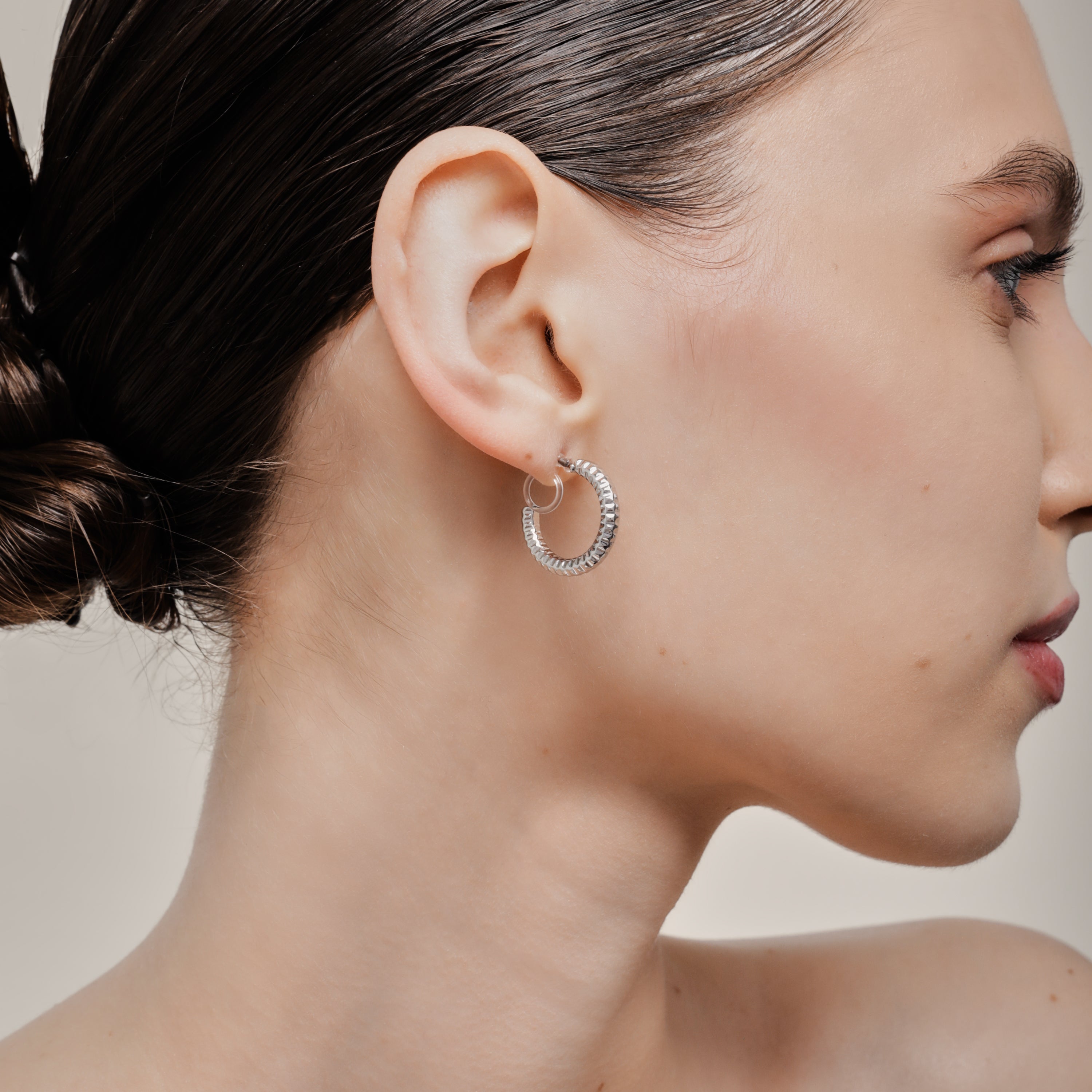 A model wearing the Ribbed Hoop Clip On Earrings in Silver. Made with a Resin Clip-On closure, these earrings provide a secure hold for 8-12 hours and are suitable for all ear sizes and types. Elevate your look with these versatile and long-lasting earrings. Set includes one pair.