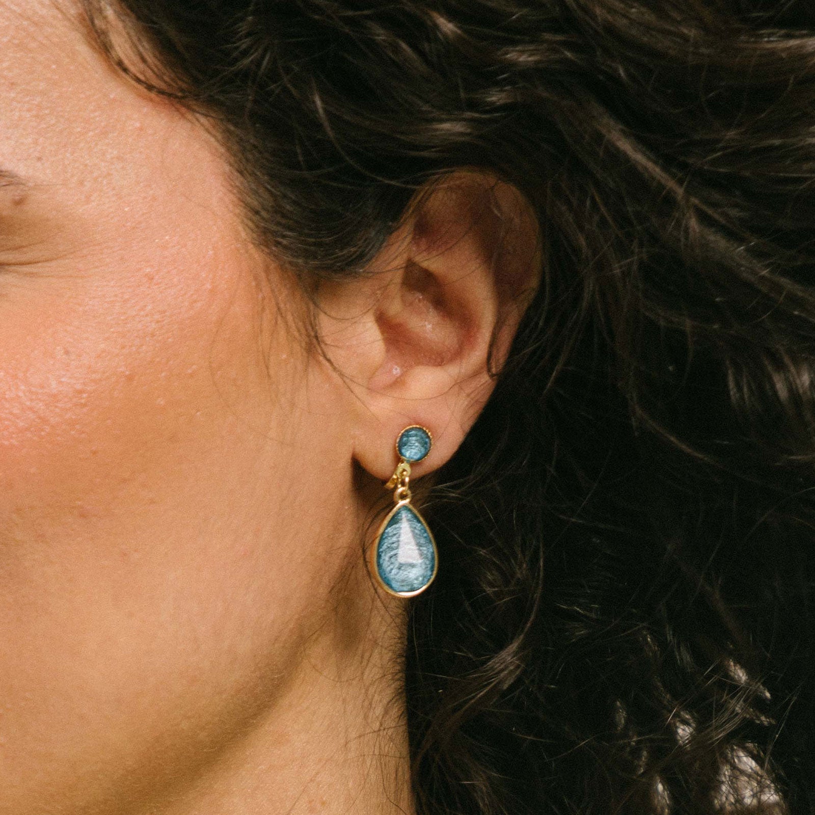 A model wearing the Sabrina Blue Tear Drop Clip-On Earrings feature a padded clip closure, making them ideal for all ear types. The earrings offer secure hold for up to 8-12 hours and are constructed of resin and gold tone plated zinc alloy. For extra comfort, the earrings come with removable rubber padding and cannot be adjusted for size. Please note, one package contains one pair of earrings.