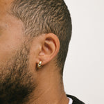 A model wearing the Small Hoop Clip On Earrings in Gold feature a mosquito coil closure for a secure hold of up to 24 hours. The earrings are designed to fit any ear type, including thick or large, sensitive, small or thin, and stretched or healing ears. They possess a medium secure hold and can be adjusted to fit the wearer's ear size, simply by gently squeezing the inner padding forward. Crafted from gold tone plated copper alloy, each package contains one pair.