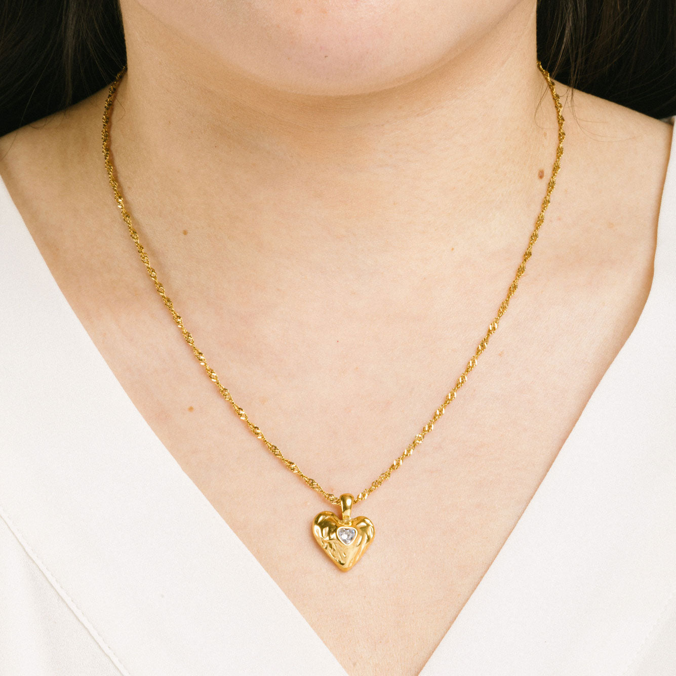 A model wearing the Textured Heart Pendant Necklace is crafted from 14K Gold Plated Stainless Steel with a Cubic Zirconia stone, and is non-tarnish, water resistant, and free from Lead, Nickel, and Cadmium. It is also adjustable and has only one necklace included.