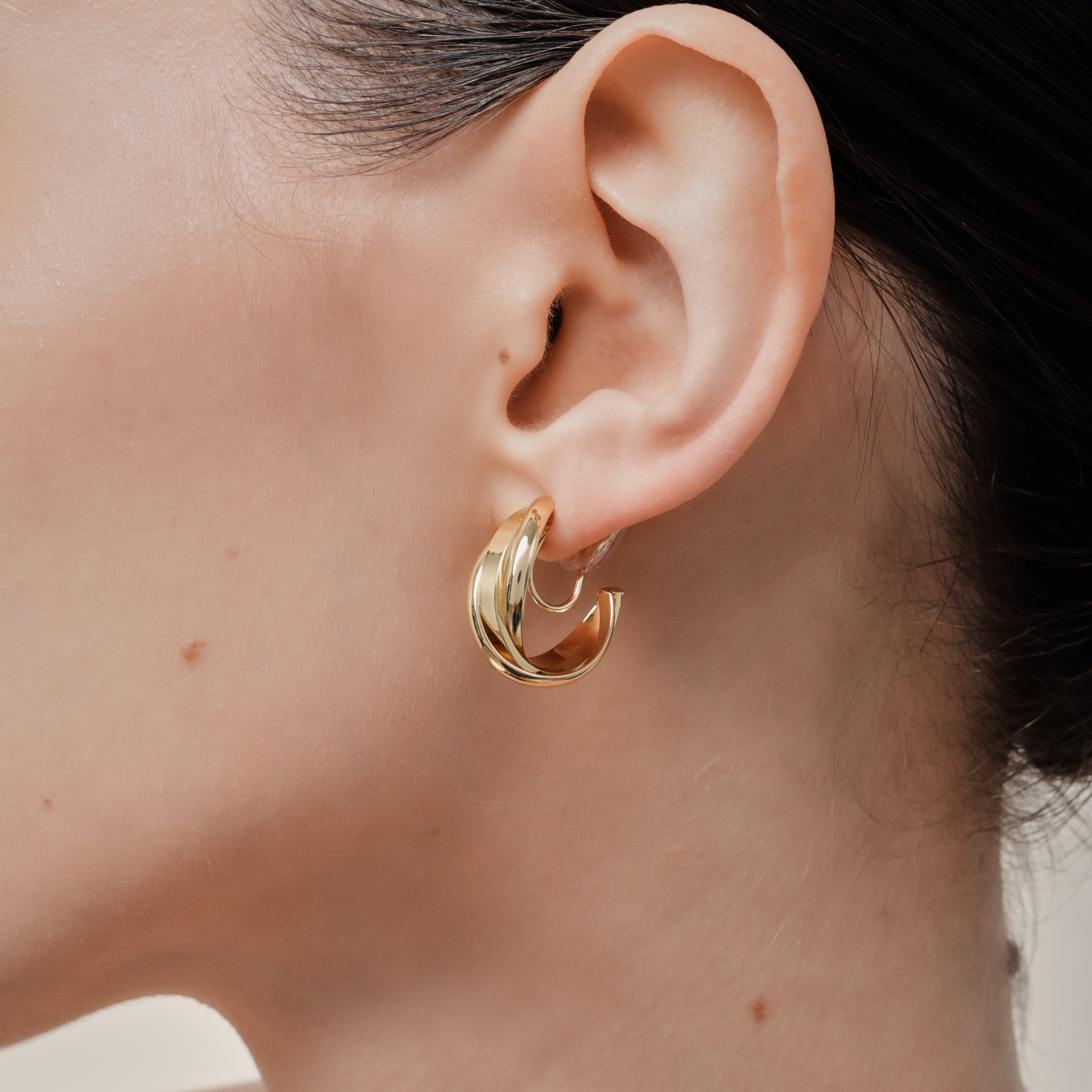 A model wearing the Trinity Hoop Clip On Earrings in Gold are perfect for those with various ear types, including thick, sensitive, small, stretched, and keloid-prone ears. Featuring a Mosquito Coil Clip-On Closure, they provide a secure hold for up to 24 hours of comfortable wear. With easily adjustable padding, these earrings are suitable for all-day wear. Please note that this item includes only one pair.
